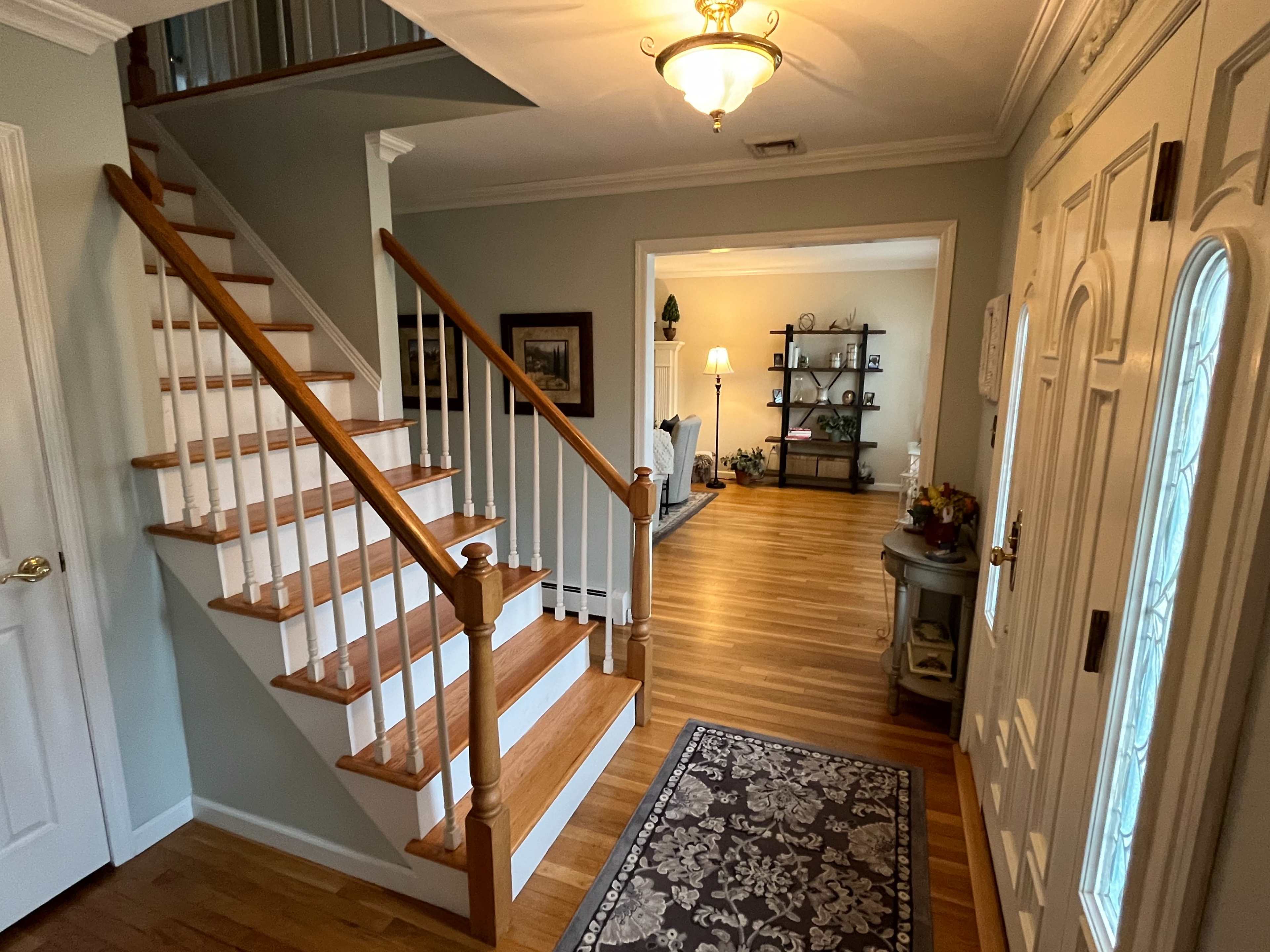 A well-lit foyer with a wooden staircase on the left, leading to an upper level, and an open view into a living area at the end.