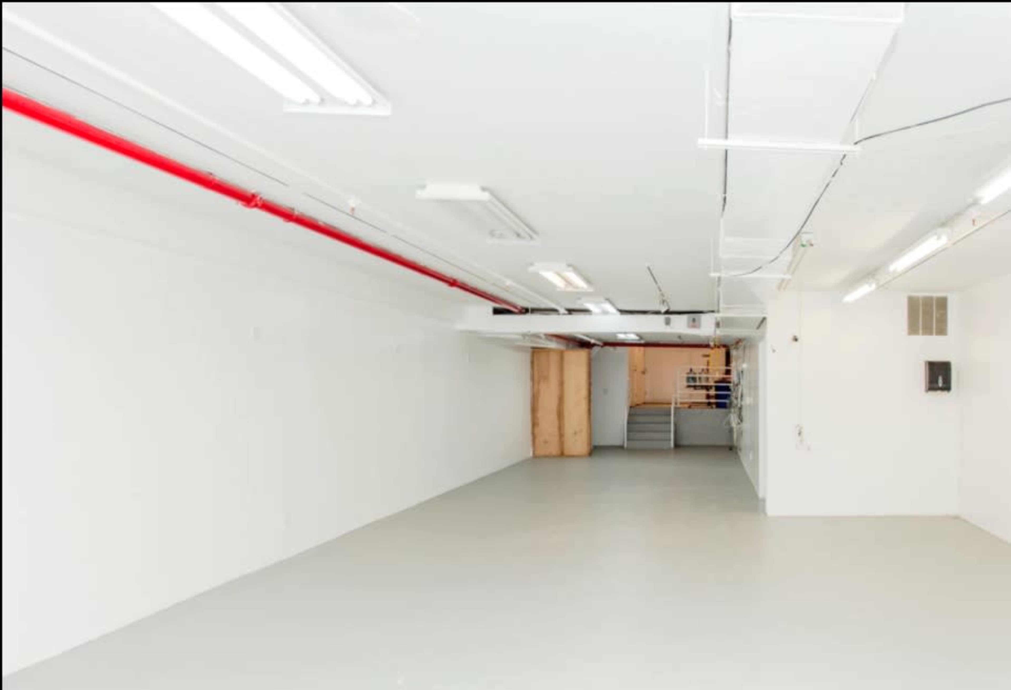 A spacious, empty room with white walls and a light gray floor, featuring red piping along the ceiling and a set of stairs in the background.