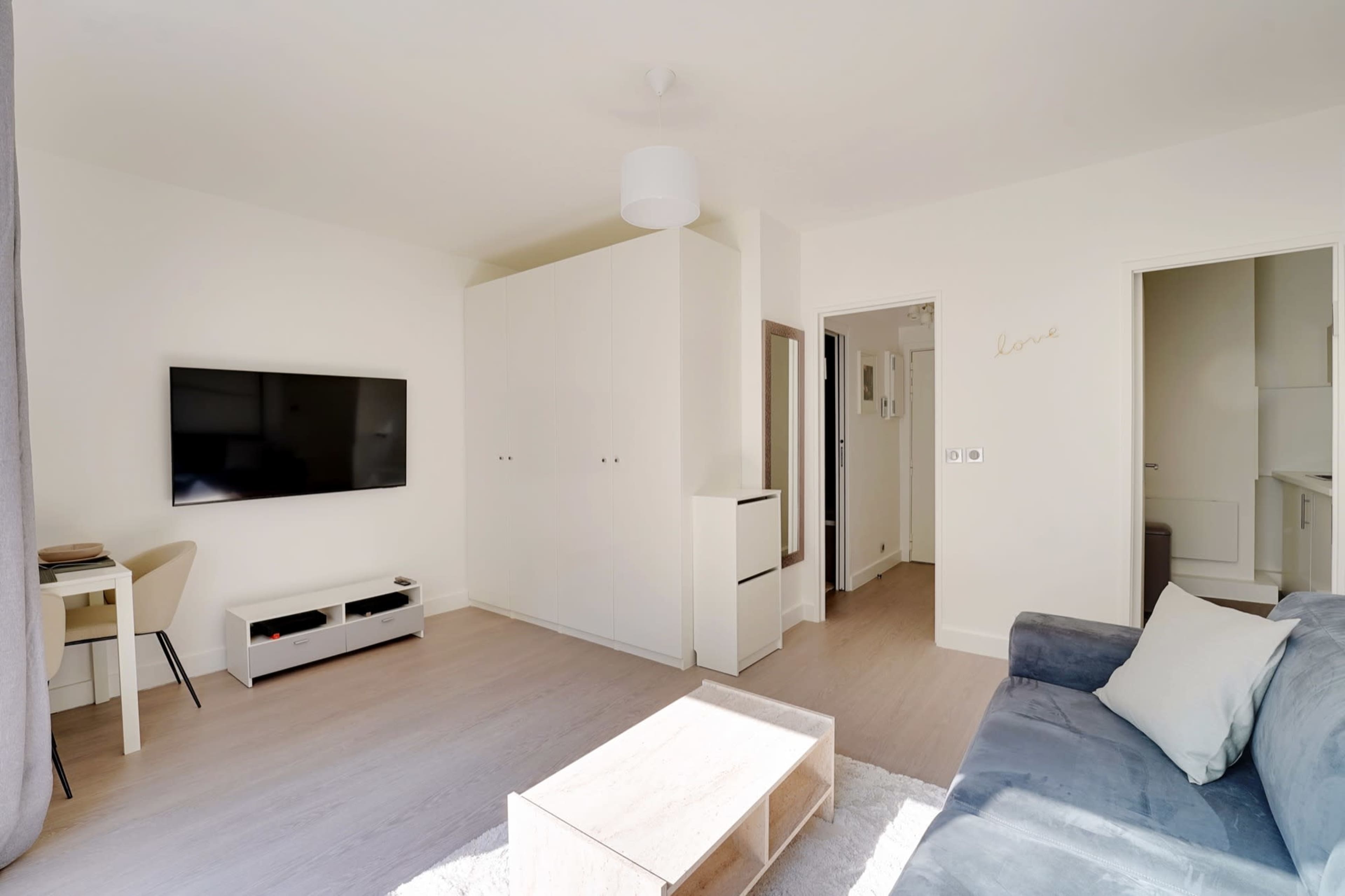 The living room features a light-colored sofa, a wooden coffee table, a wall-mounted TV, and a small dining area with a chair.