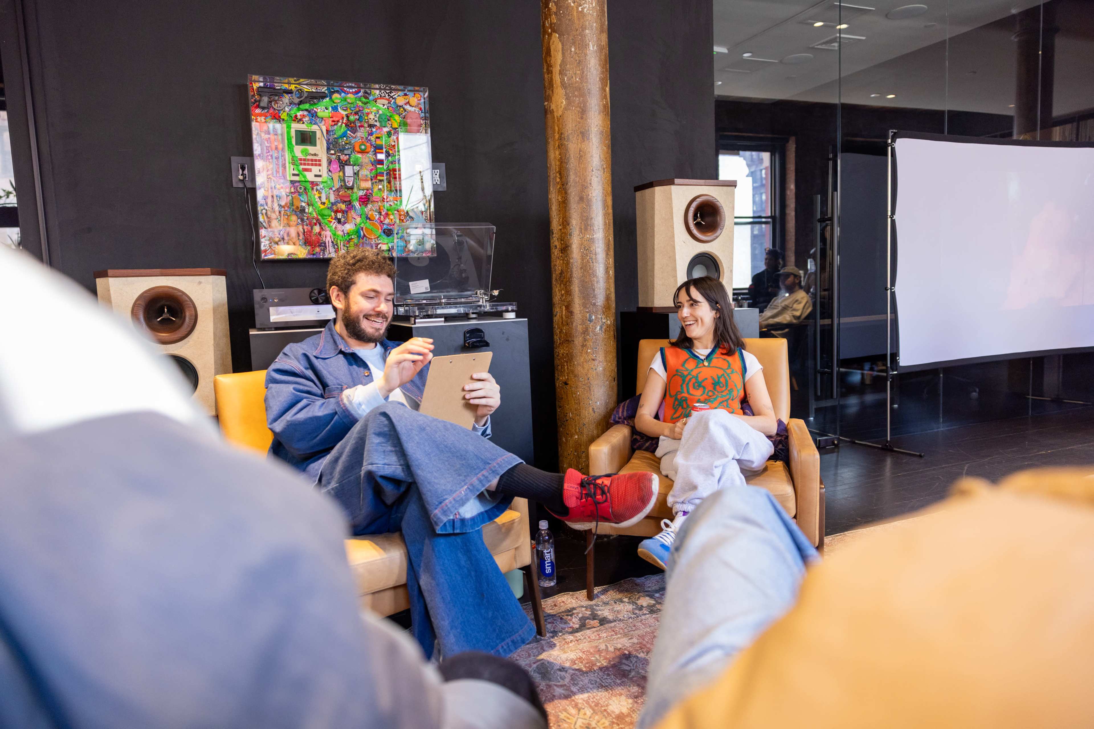 A man and a woman sit in a modern, stylish room with a large screen in the background and vintage audio equipment, engaged in conversation.