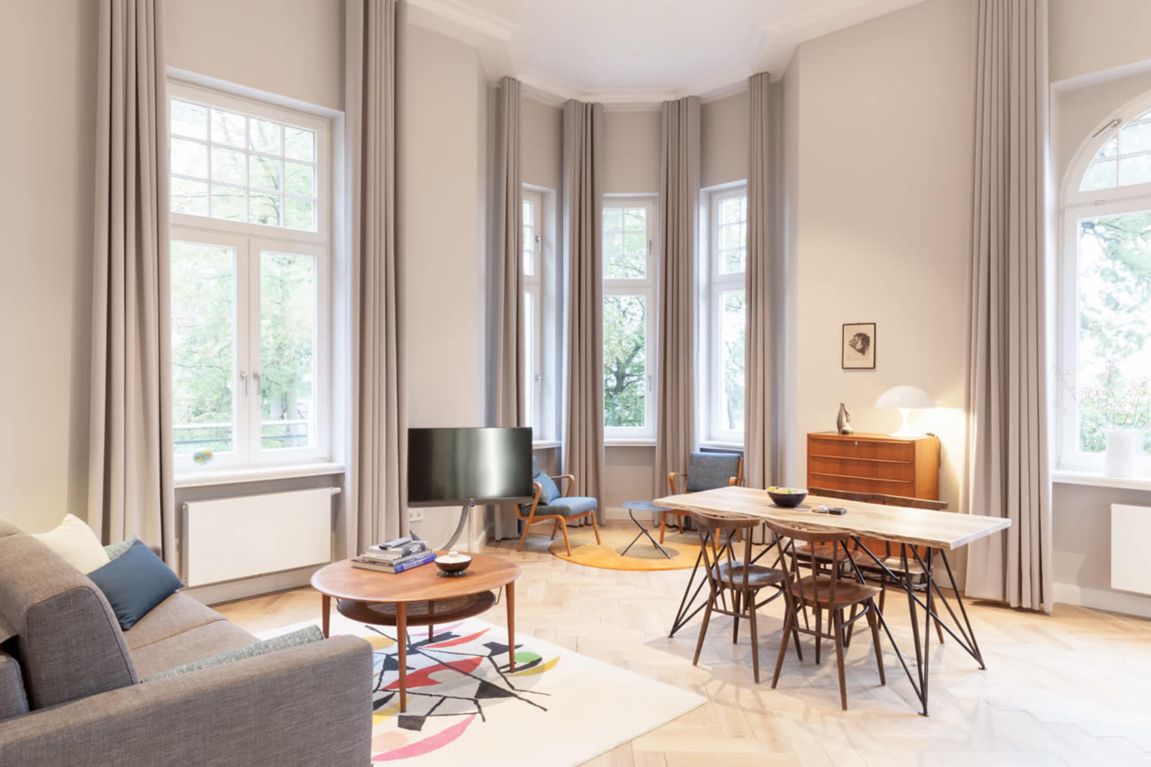 A modern living room features large windows, a comfortable sofa, a round table with chairs, and a television beside a sideboard.