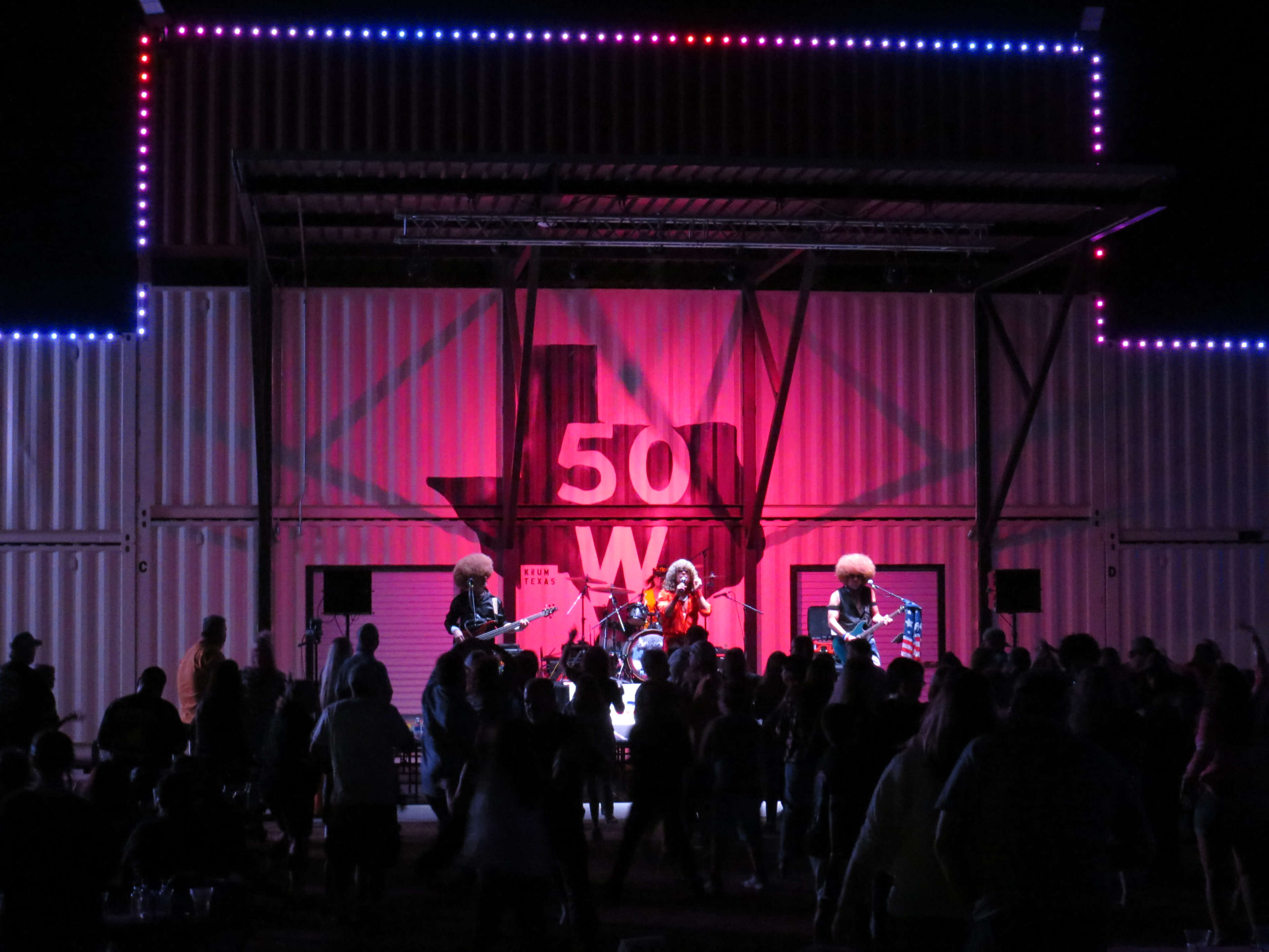 A band performs on stage in front of an audience at an outdoor venue decorated with colorful lights and a large Texas state outline.