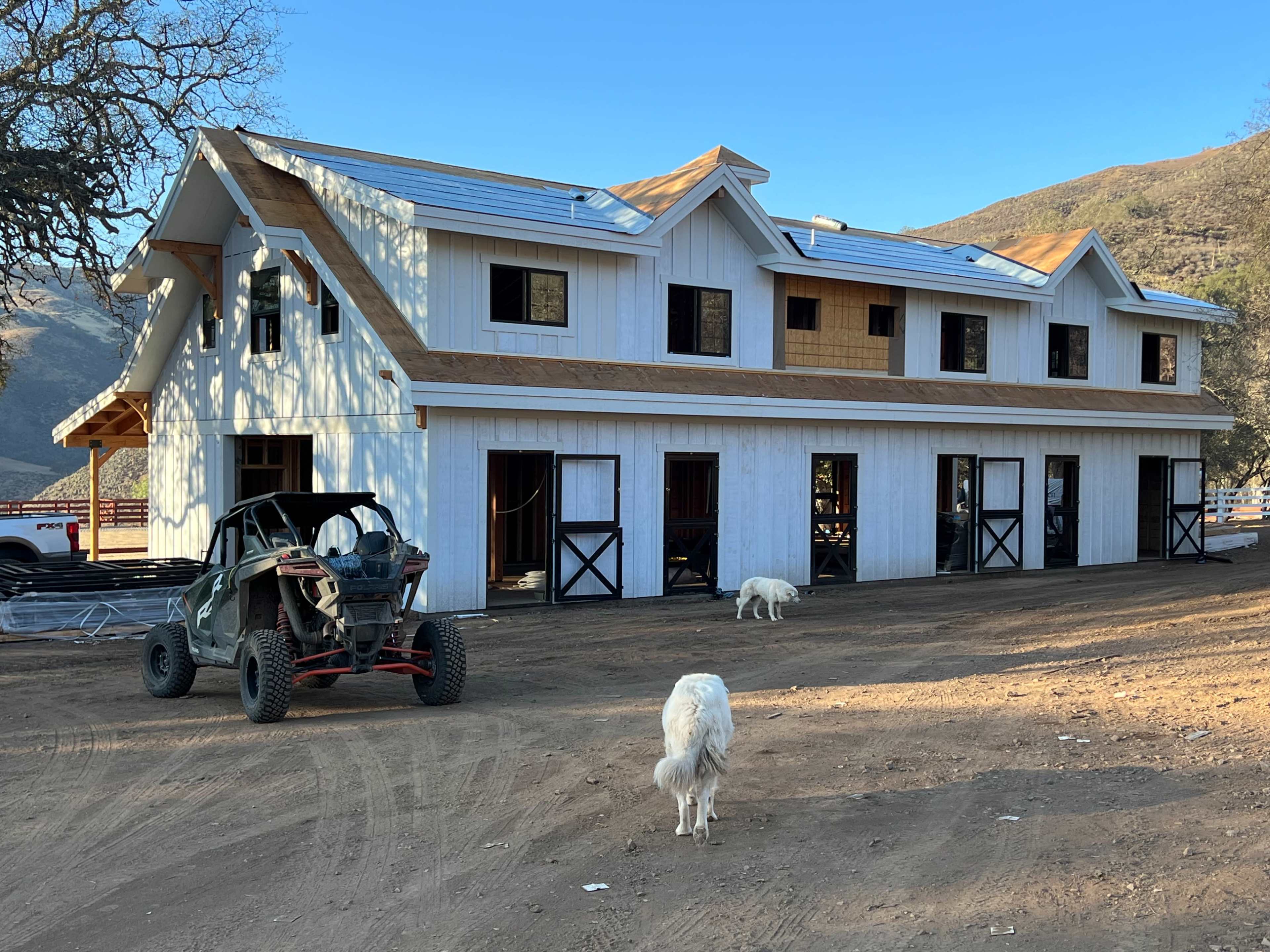 A large, partially constructed farmhouse with a white exterior and multiple windows stands in a dirt area, accompanied by a utility vehicle and two goats.