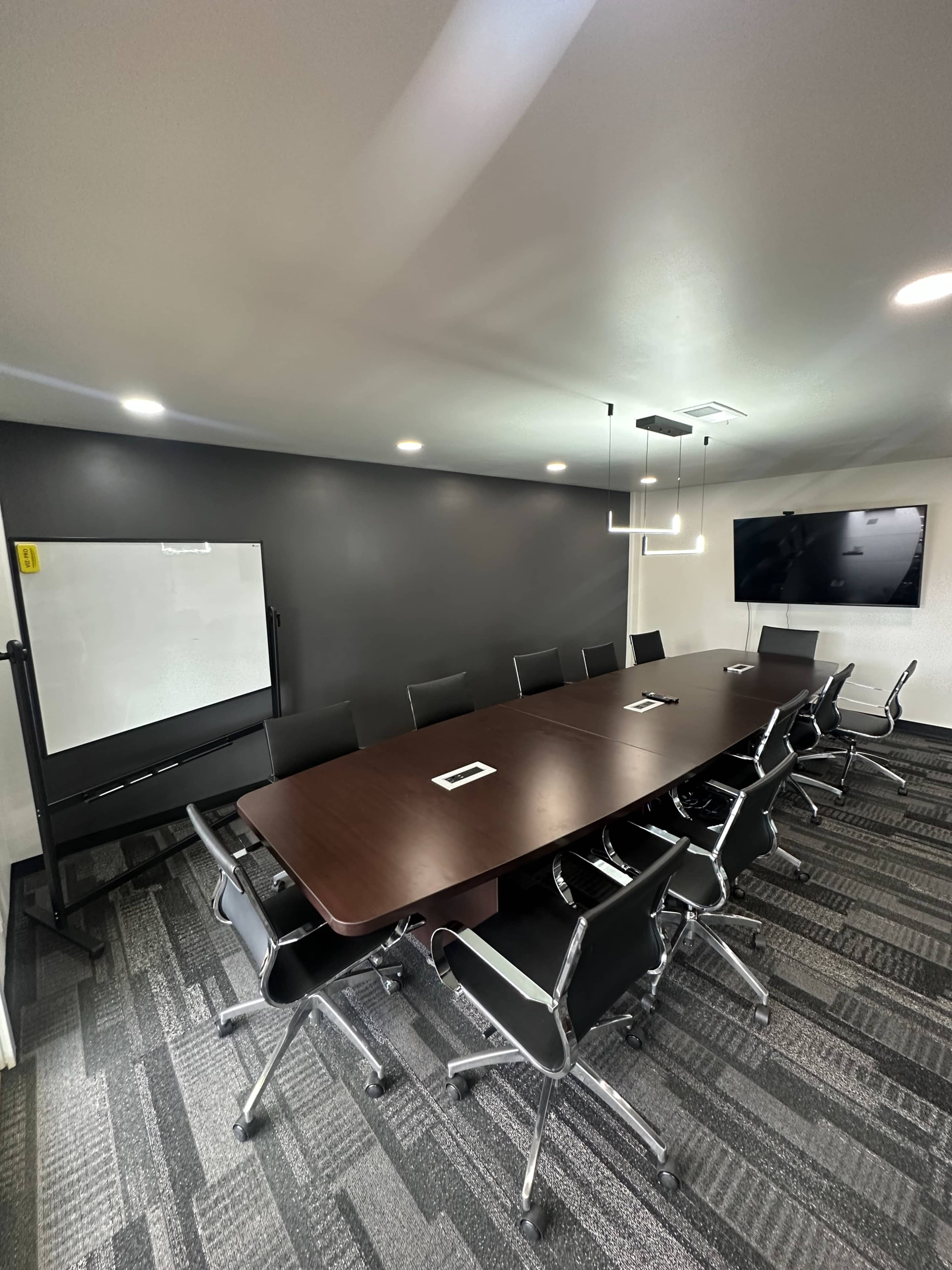 A modern conference room featuring a long wooden table surrounded by black chairs, with a whiteboard and a large screen on the walls.