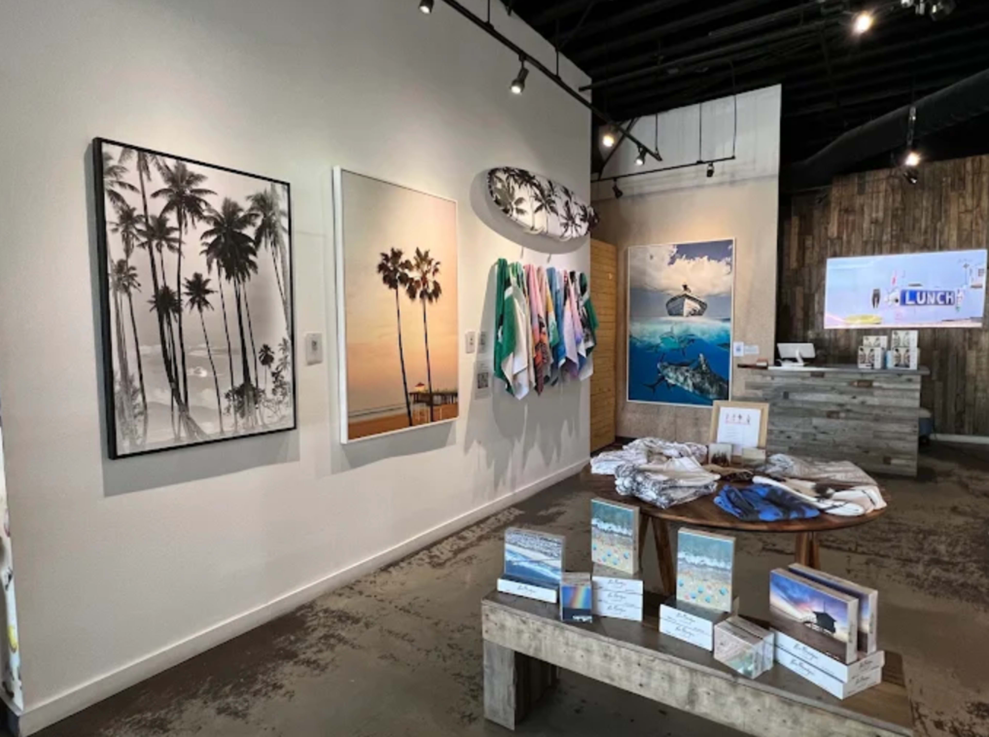 The image shows the interior of a retail space featuring artwork on the walls, clothing displays hanging, and a merchandise table in the center.