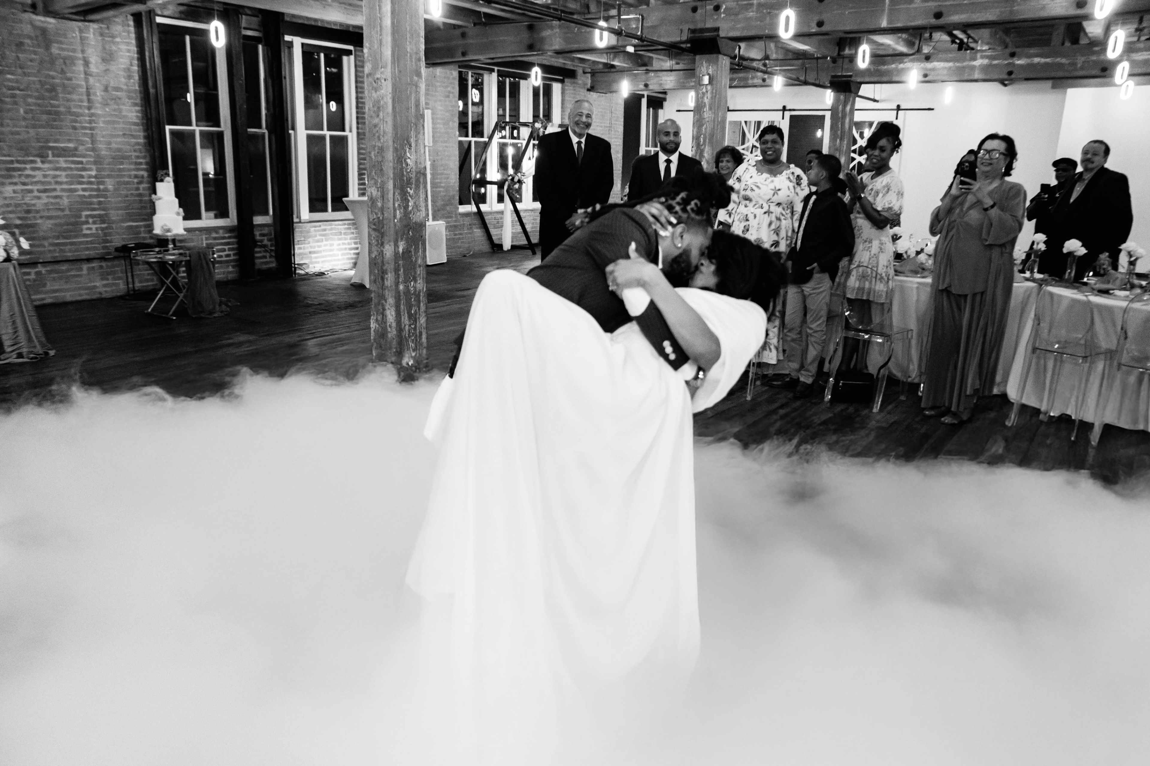 A couple performs a joyful first dance amidst a cloud of fog, with many guests watching and celebrating in a stylish venue.
