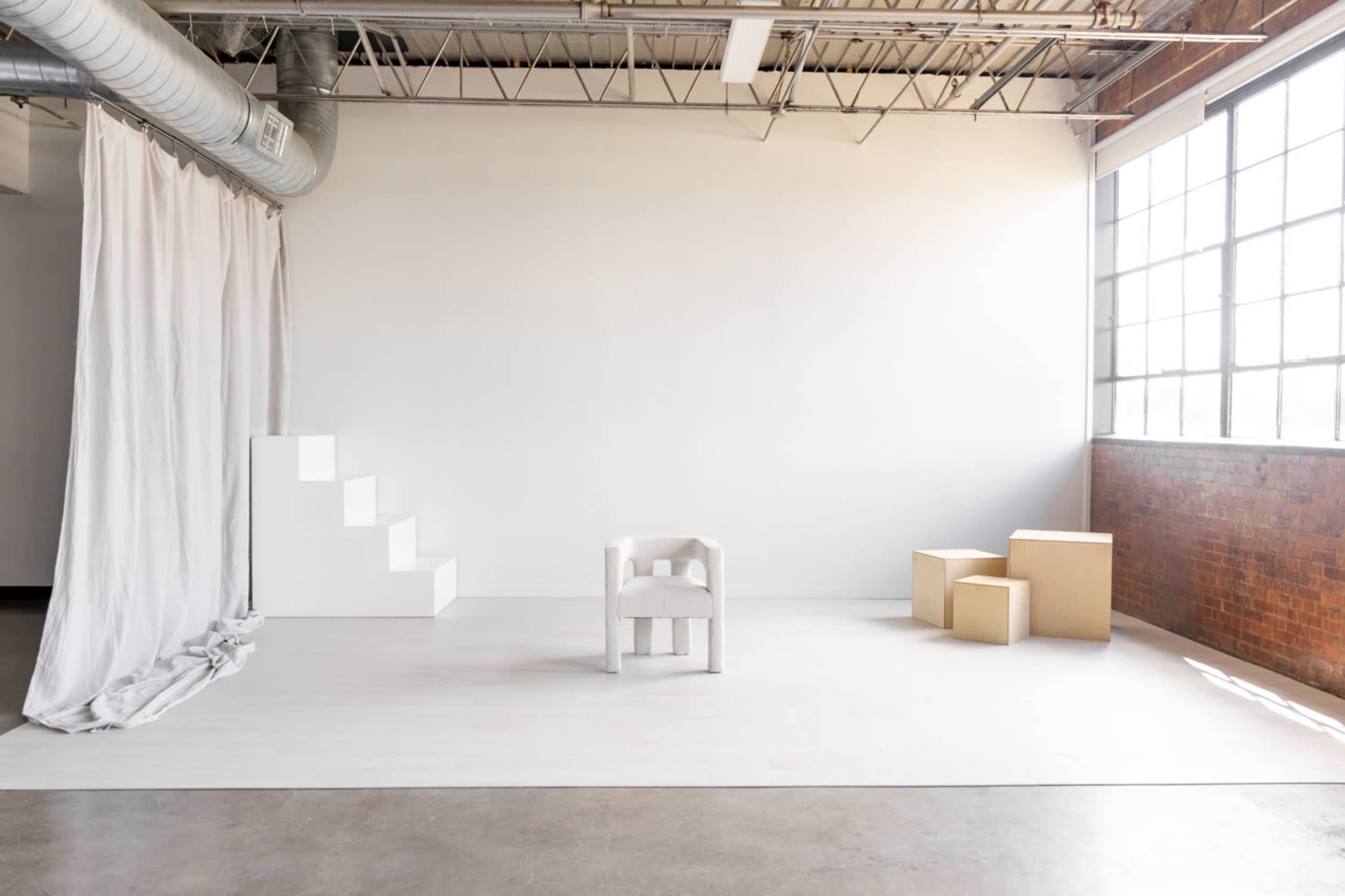 The image shows a minimalist interior space featuring white walls, a set of stairs, a transparent chair, and several wooden boxes.