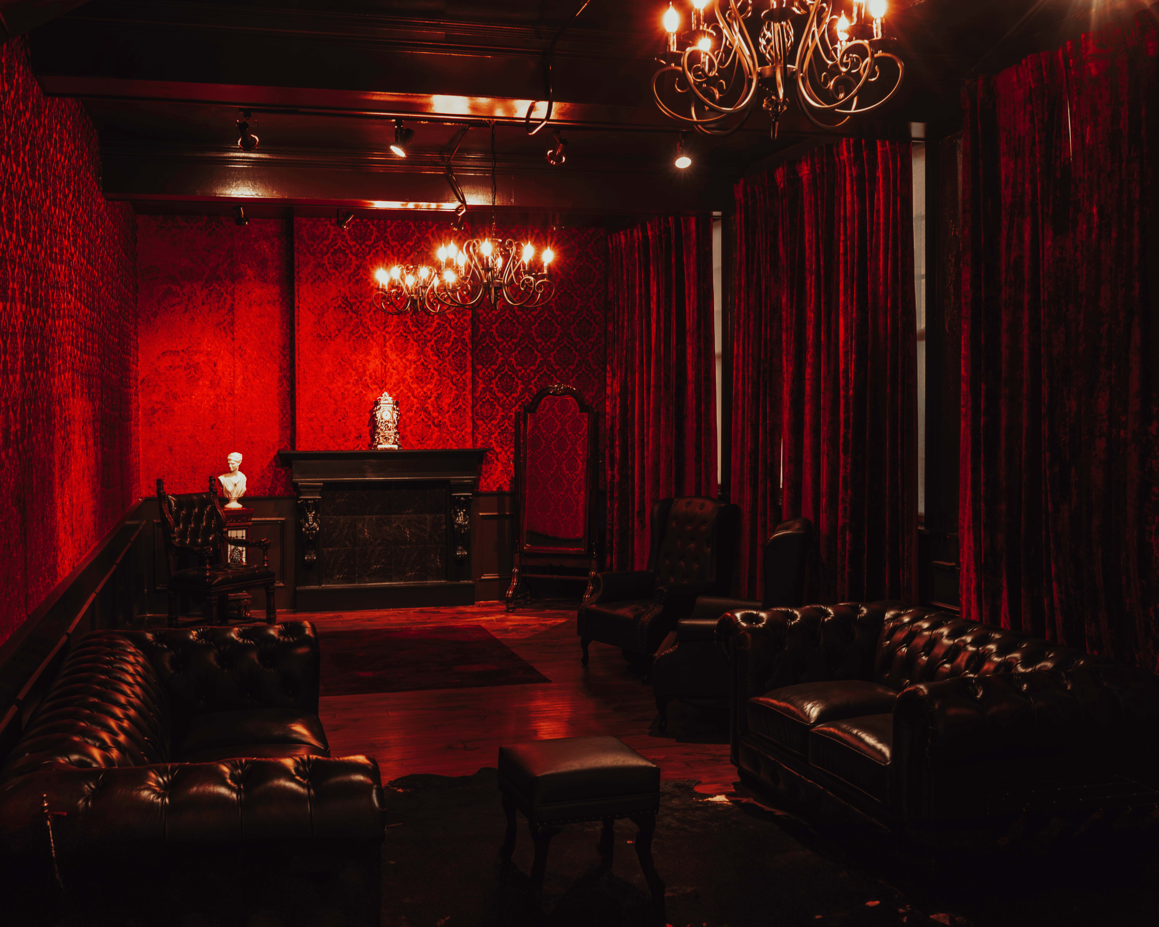 The room features red-patterned wallpaper, dark leather furnishings, and a central chandelier illuminating a fireplace and a decorative bust.