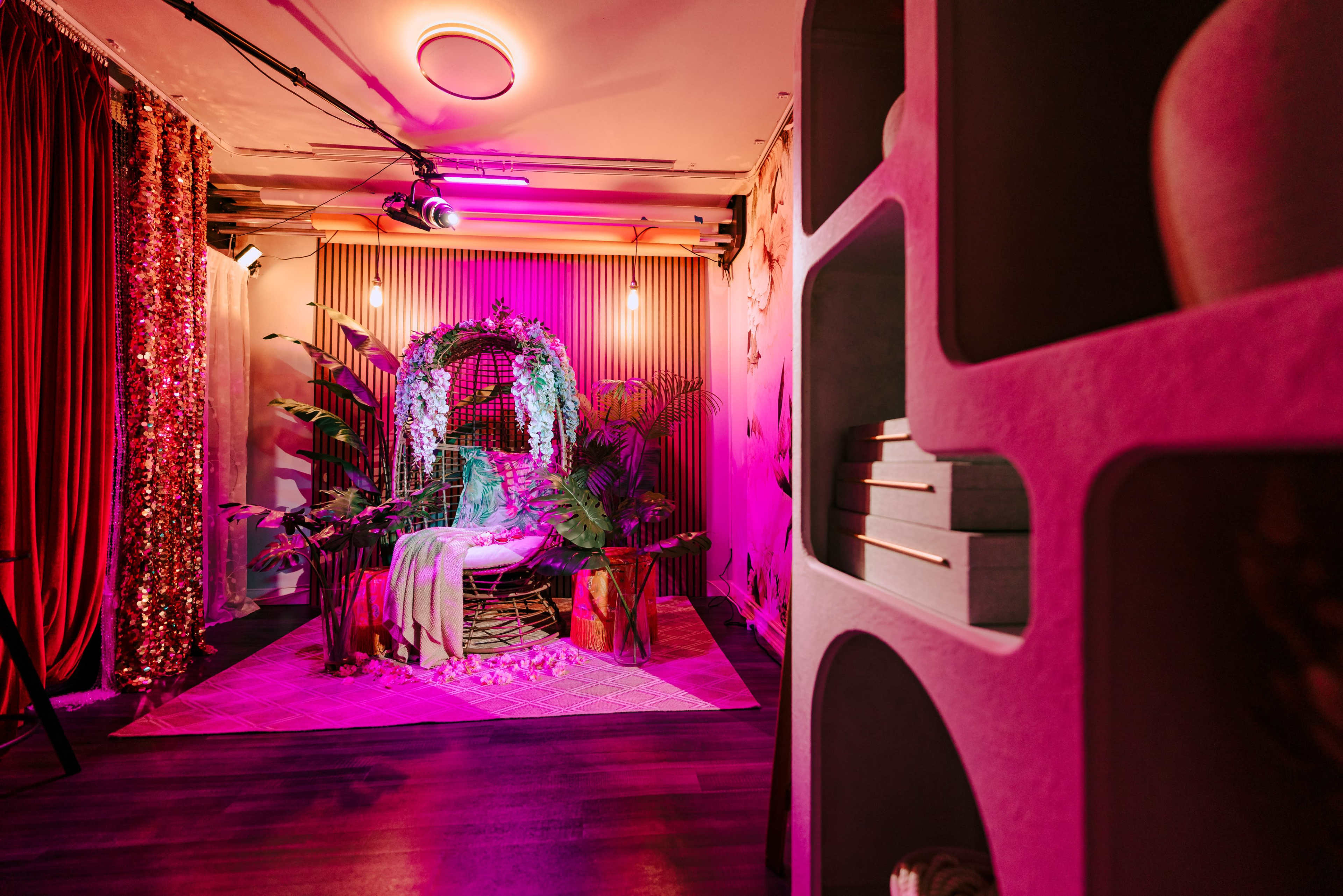 The image shows a cozy, decorated indoor space featuring a seating area surrounded by plants, bright lighting, and colorful drapery.
