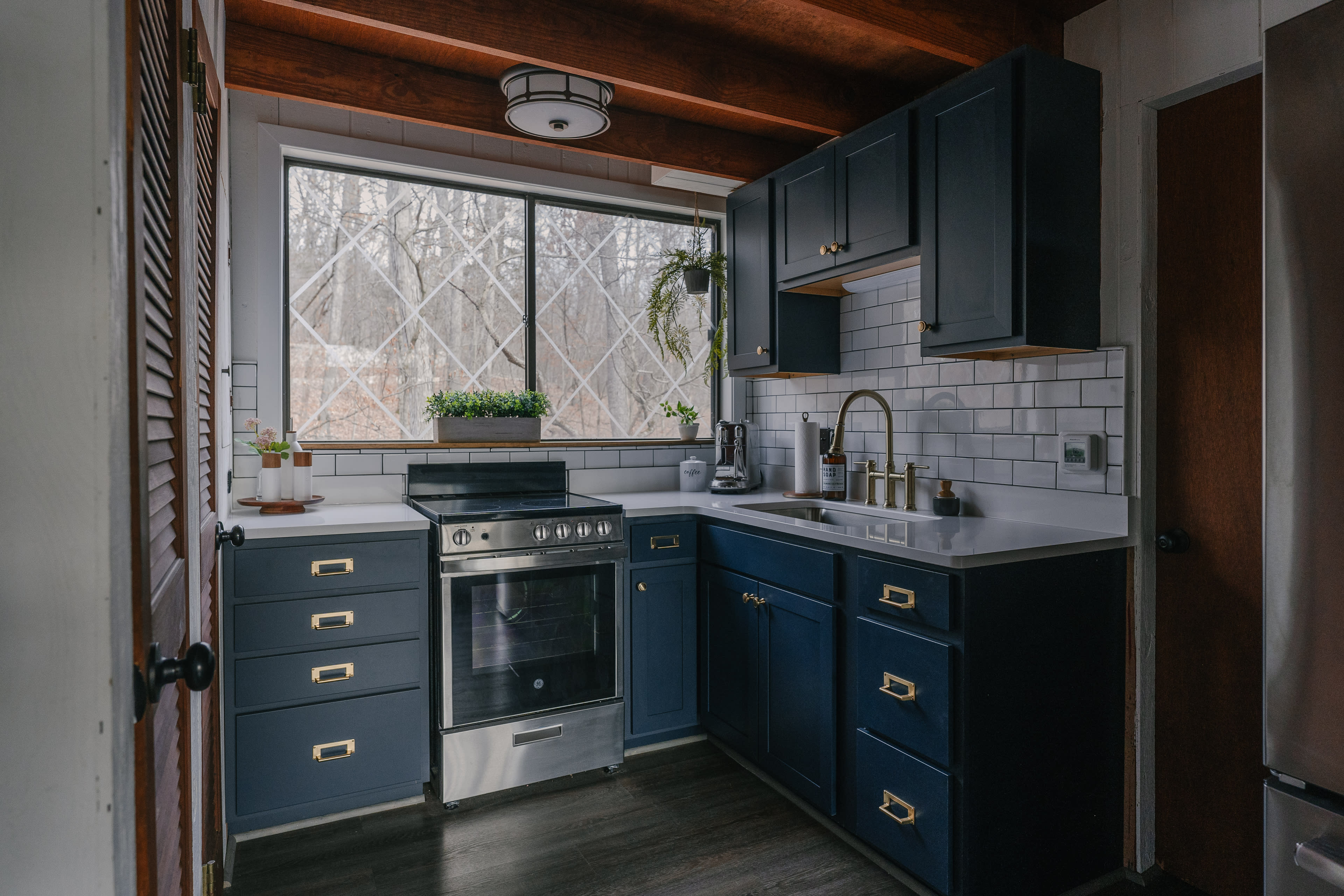 The image shows a modern kitchen with dark blue cabinetry, a stainless steel stove, and a window featuring a view of trees outside.