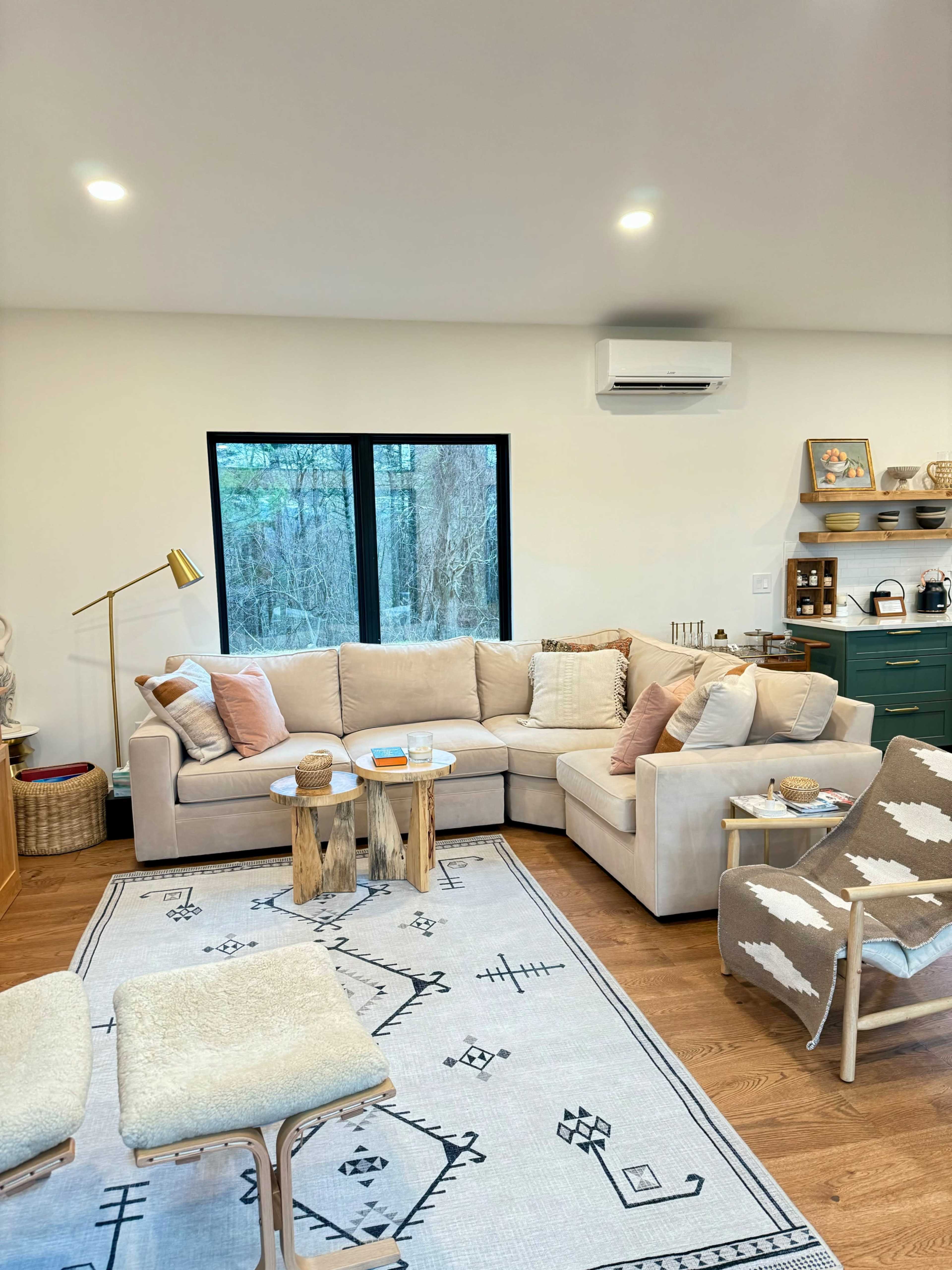 The living room features a large, light-colored sectional sofa with decorative pillows, a wooden coffee table made of tree stumps, and a patterned rug, all arranged around a window that looks out onto a wooded area.