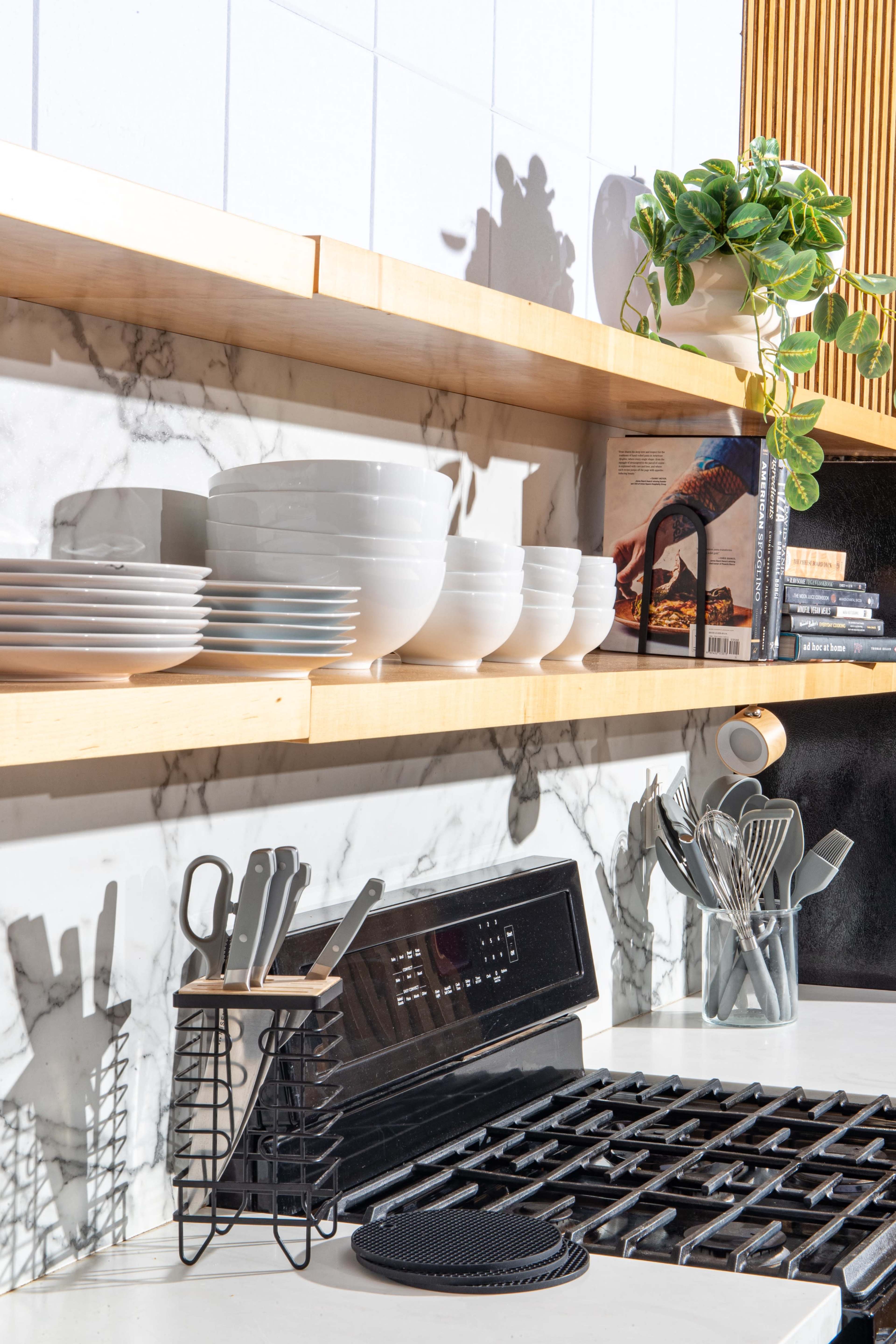 A modern kitchen countertop features stacked white bowls and plates on wooden shelves, a knife holder, and various cooking utensils alongside a stovetop.