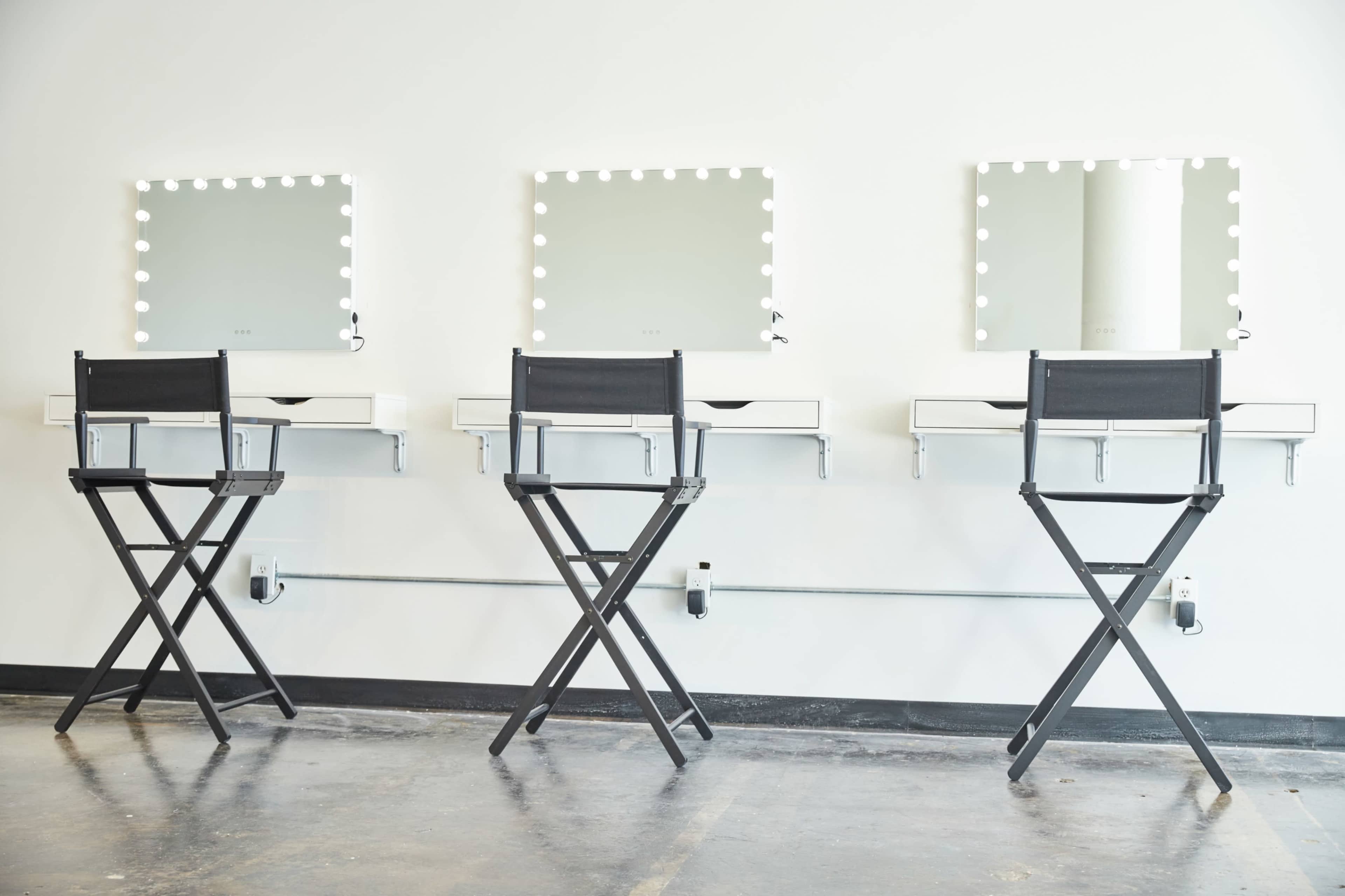Three makeup stations with mirrors and lights, each accompanied by a black folding chair.