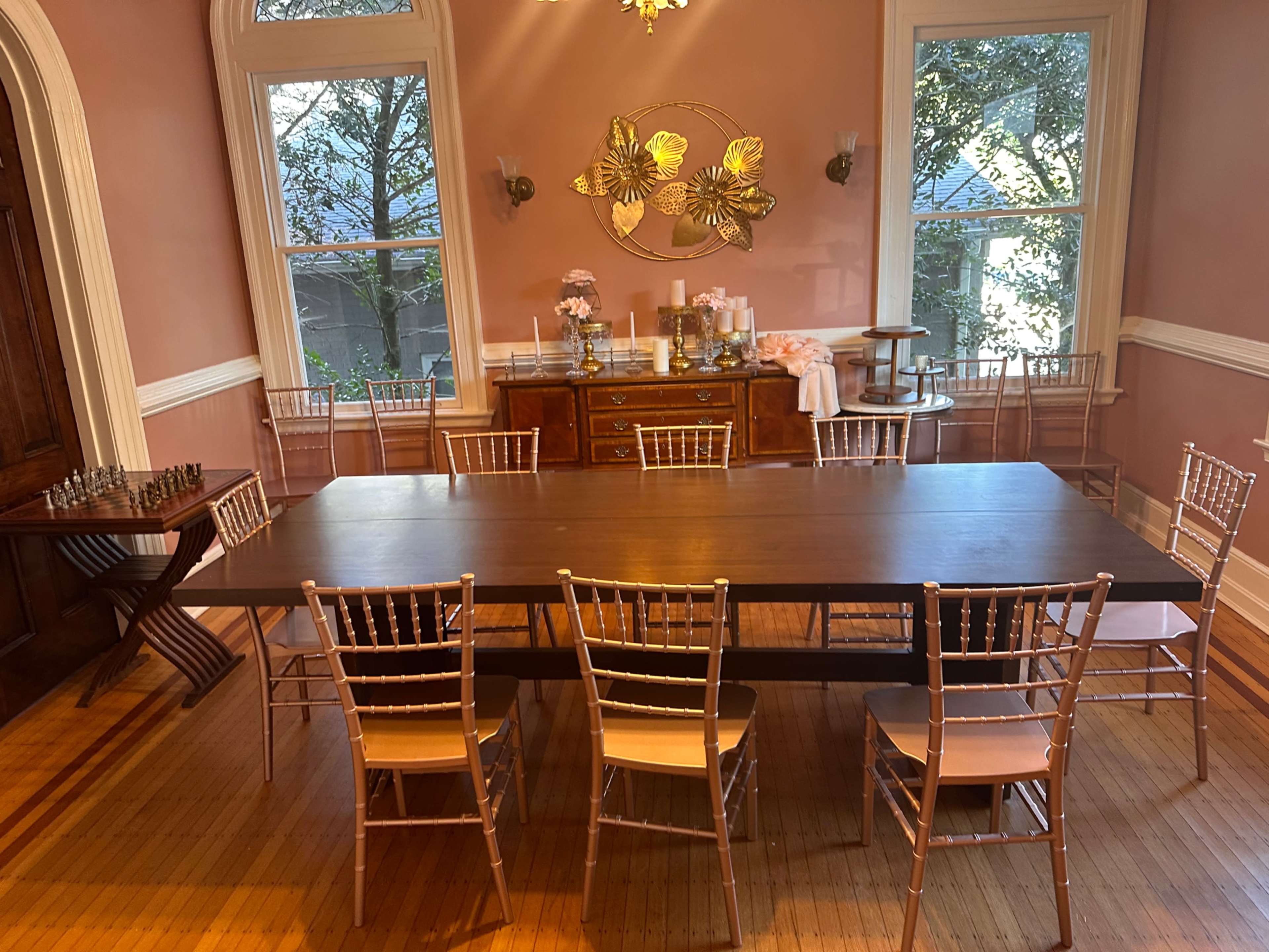 A long wooden dining table surrounded by gold chairs is set in a room with large windows and pink walls.