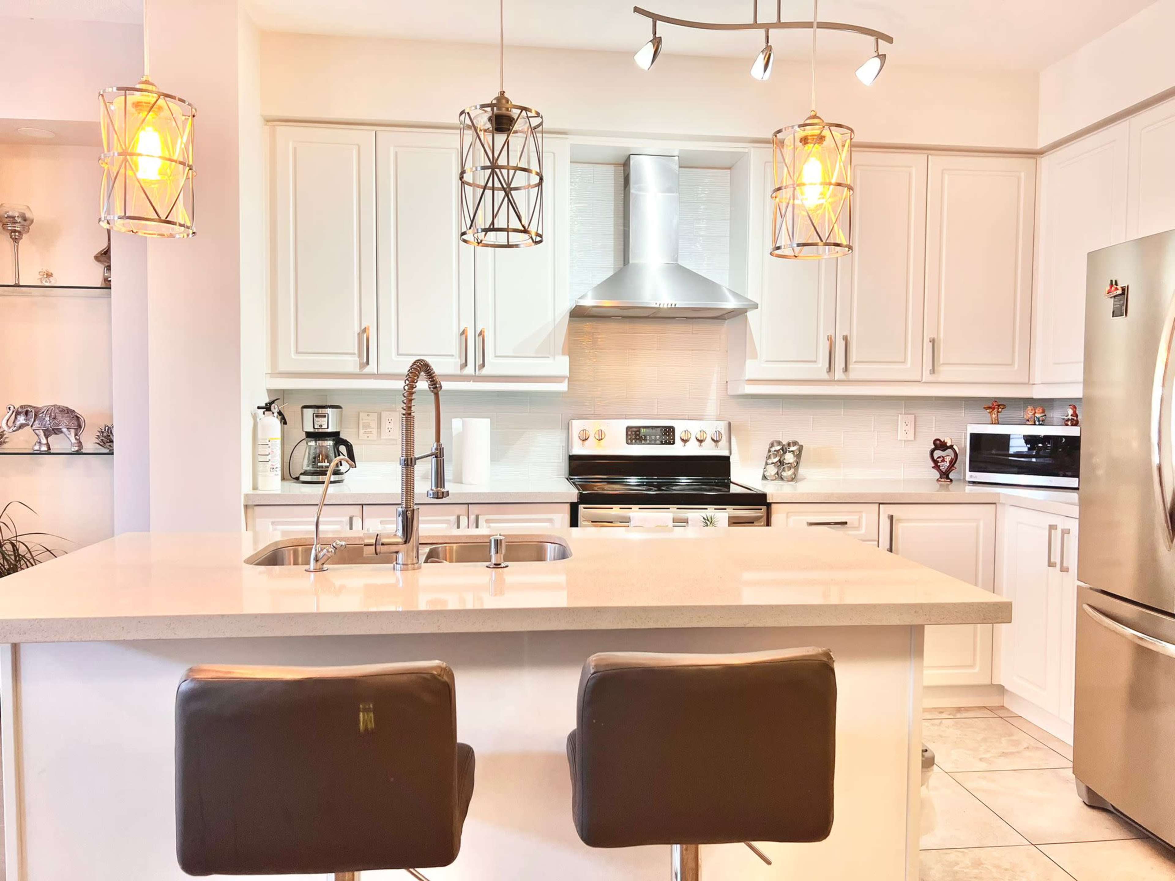 A modern kitchen featuring a central island with two black bar stools, overhead pendant lighting, and sleek white cabinetry.
