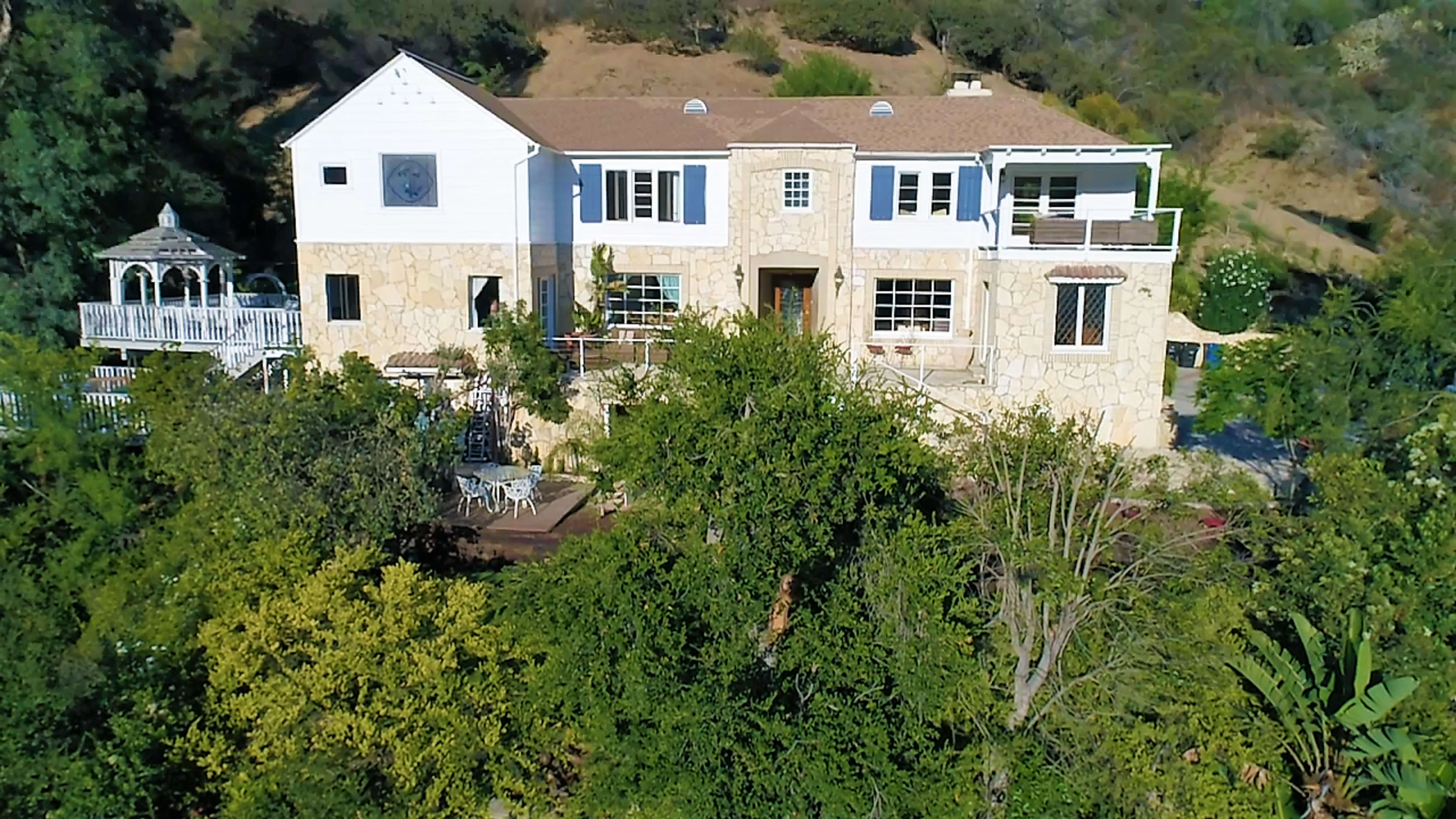 A two-story stone and white house surrounded by greenery on a hillside.