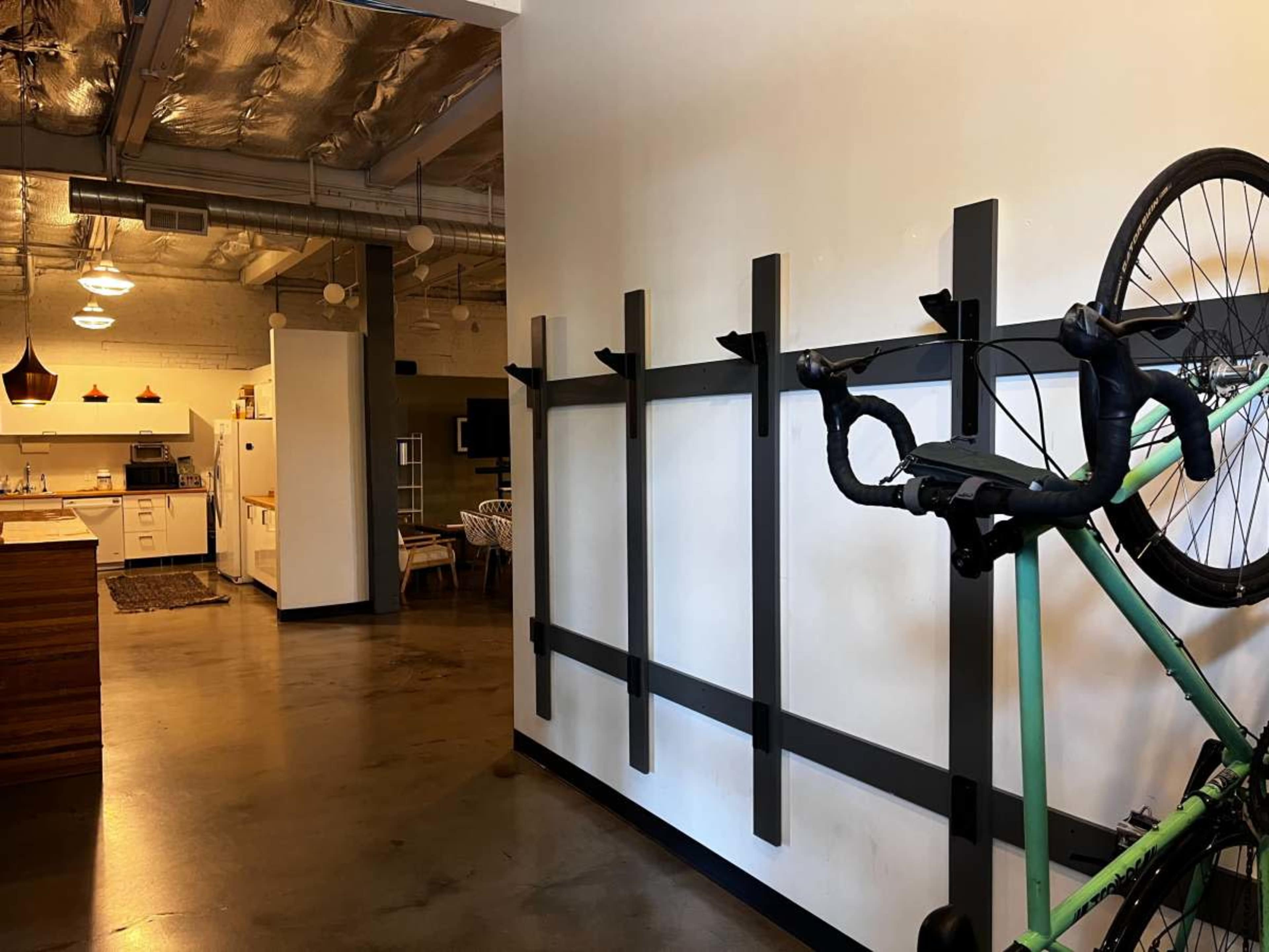 A green bicycle is hung on a wall-mounted rack in a modern interior space that features a kitchen area in the background.