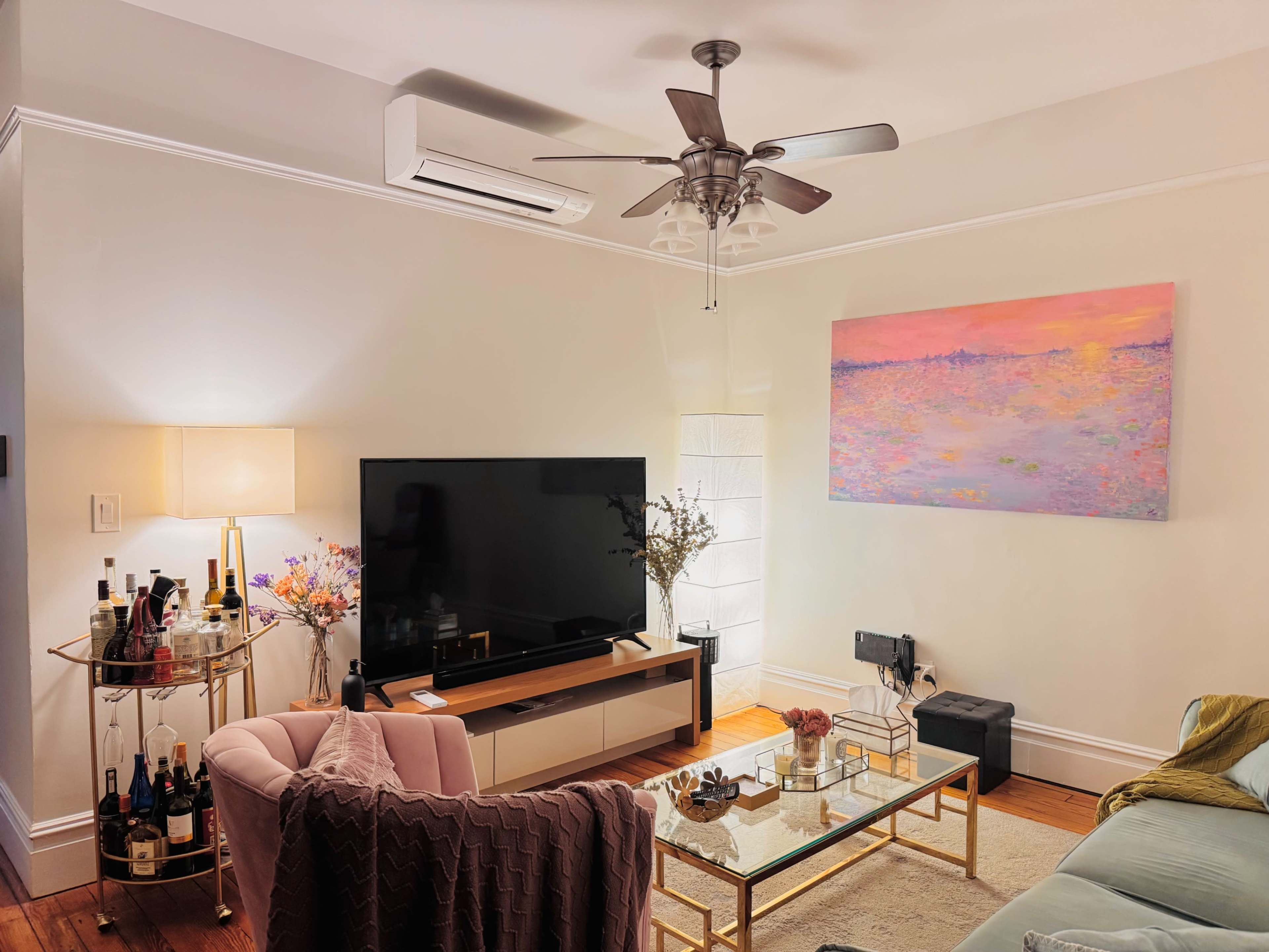 The image shows a cozy living room with a television, a glass coffee table, a pink armchair, and a wall-mounted air conditioning unit.