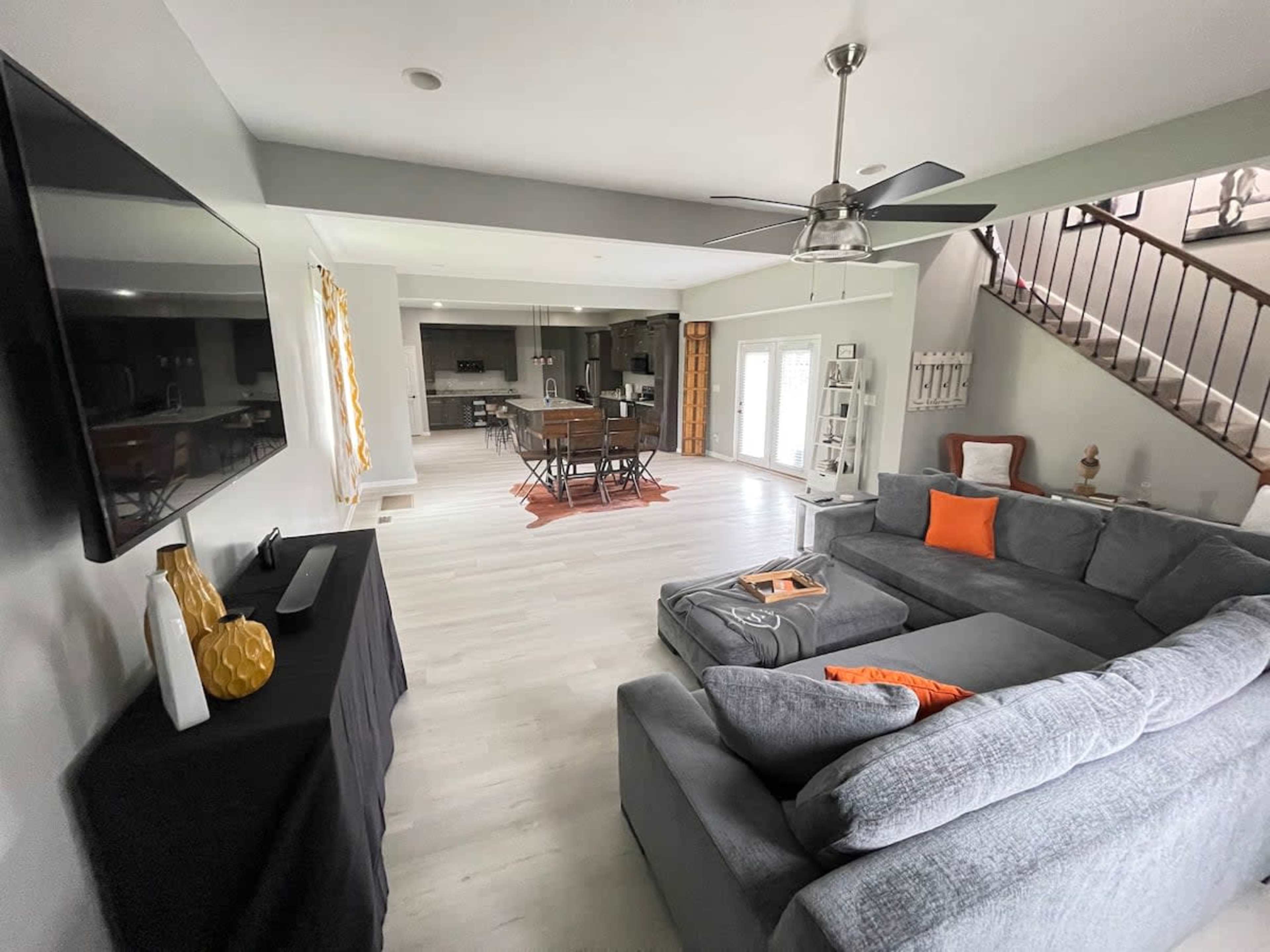 A spacious living area with a gray sectional sofa, a flat-screen TV mounted on the wall, and a dining area visible in the background.