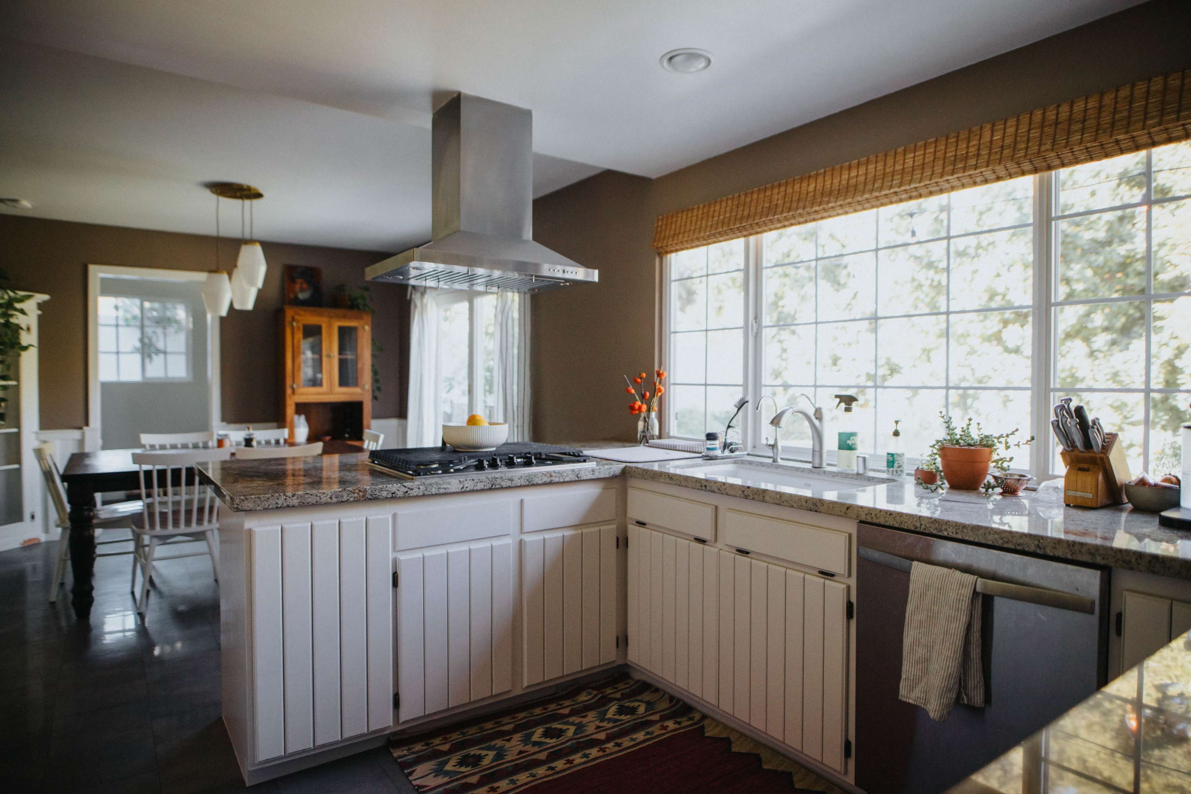 The image shows a modern kitchen with a large window, granite countertops, a central island with a stovetop, and potted plants on the countertop.