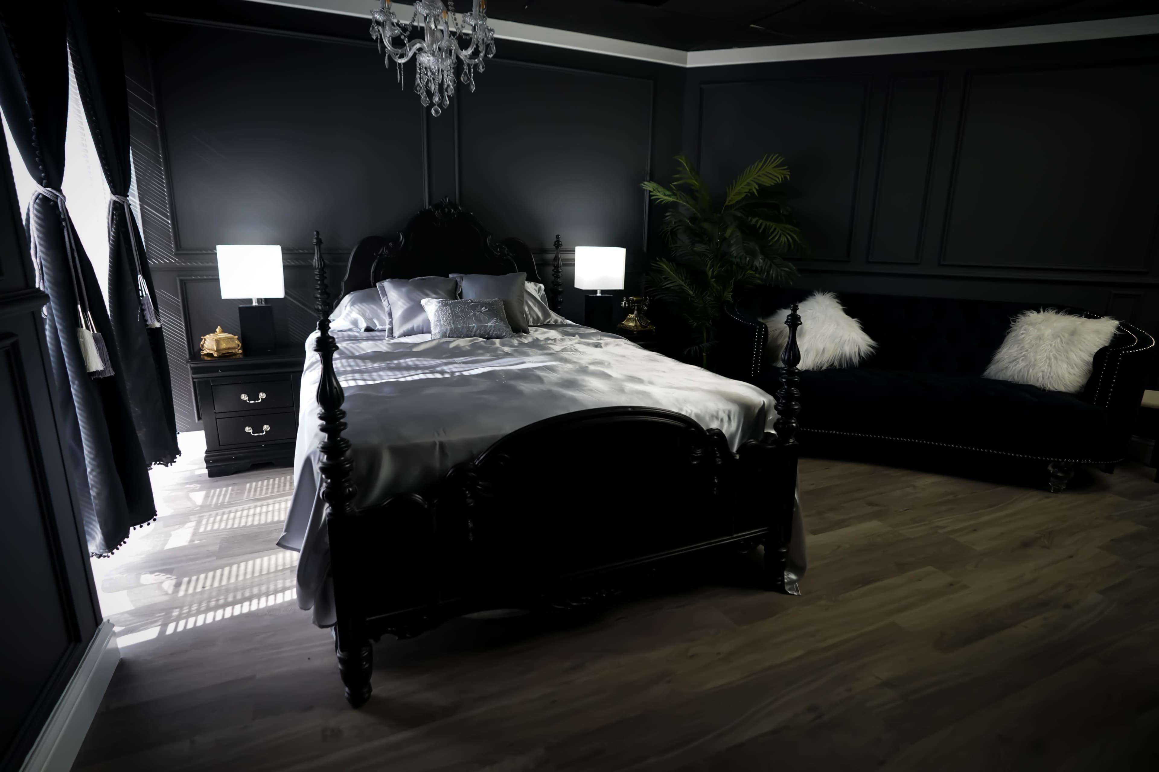 A darkly themed bedroom featuring a large black bed with a chandelier overhead, flanked by two lamps and a plush sofa.