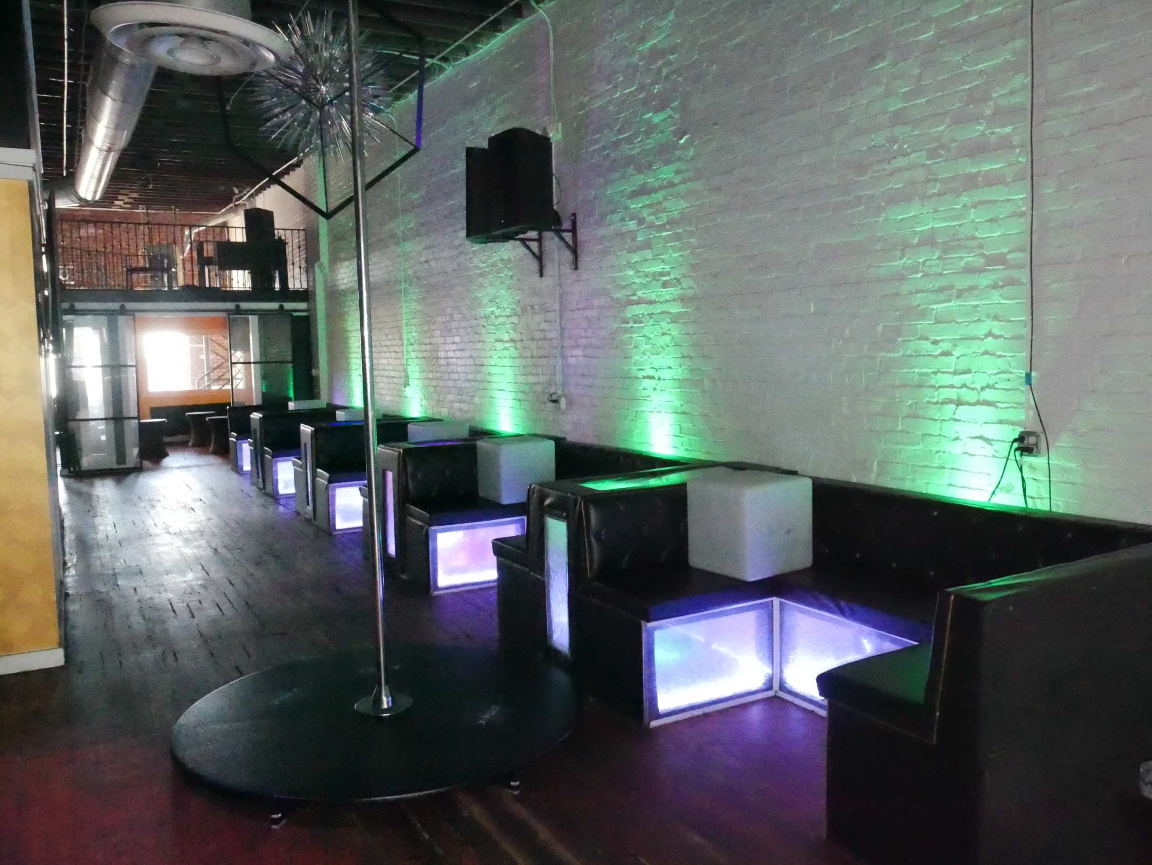 The interior of a nightclub features a row of illuminated seating areas and a central dance pole, with green lighting highlighting the space.