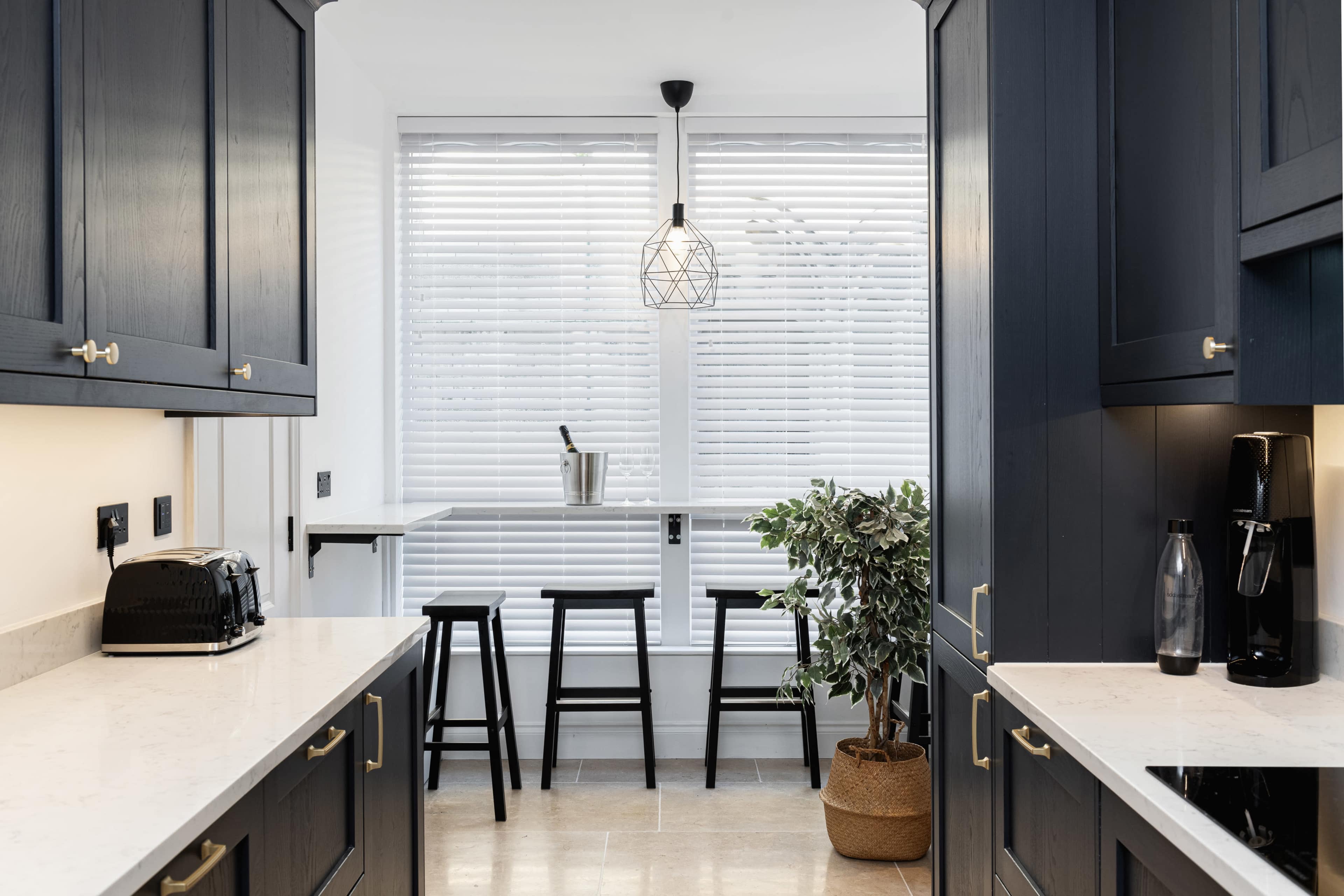 A modern kitchen with dark cabinetry, a marble countertop, and a small dining area featuring black stools and a plant.