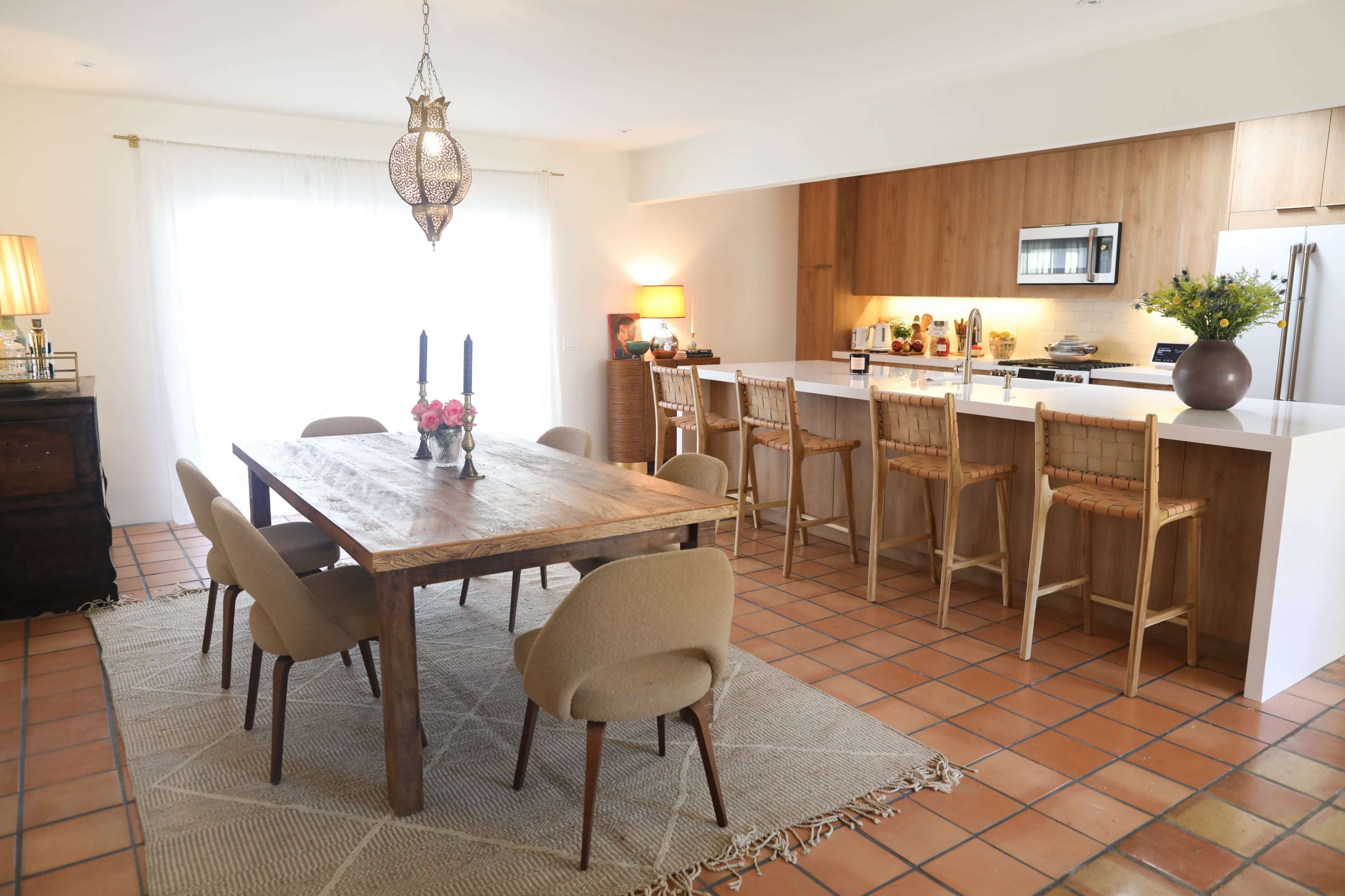 A bright dining area featuring a wooden table with chairs, a kitchen area with bar seating, and large windows allowing natural light to enter.