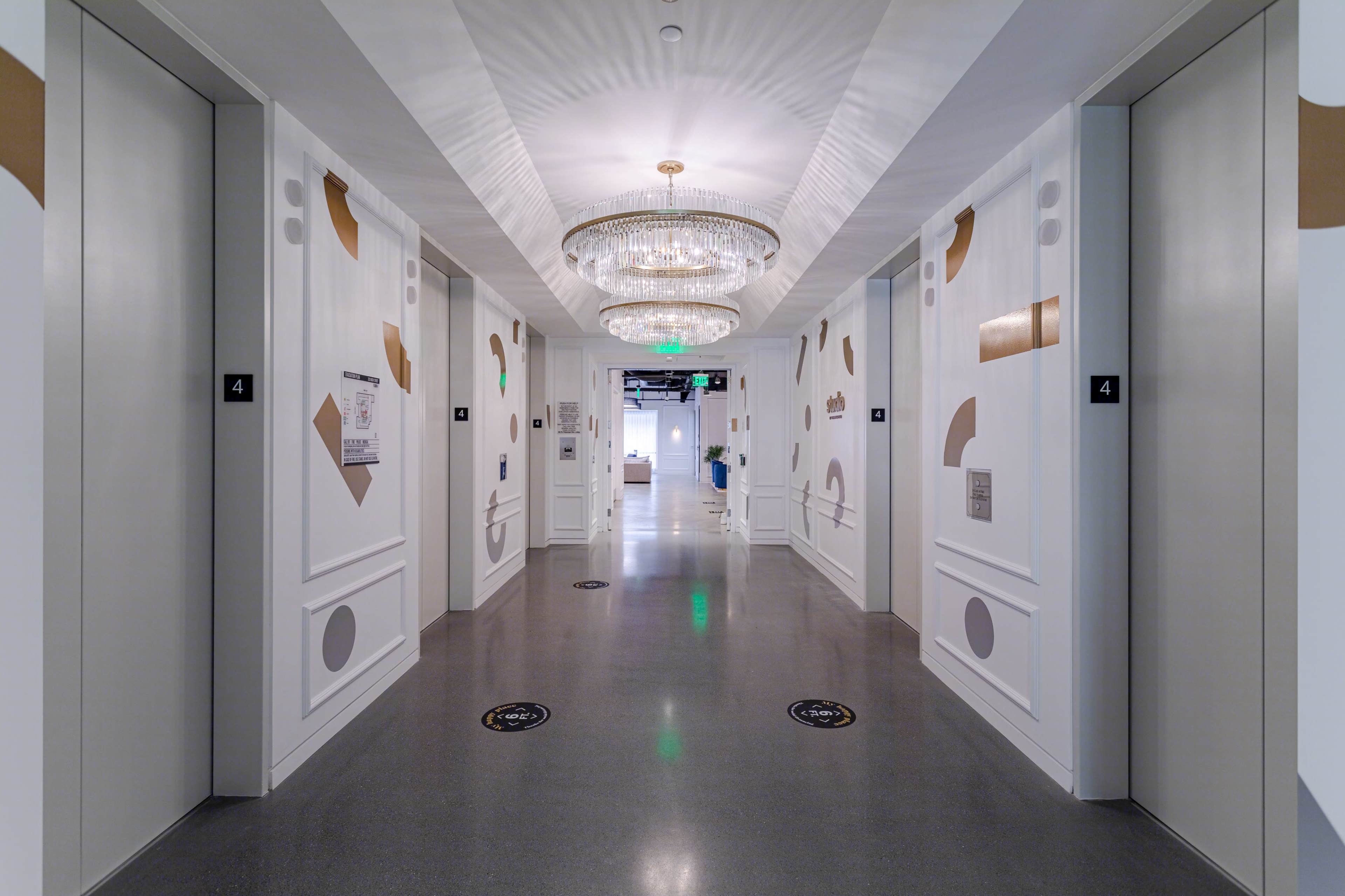 A wide hallway features elevator doors on either side, with modern wall decor and a chandelier overhead.