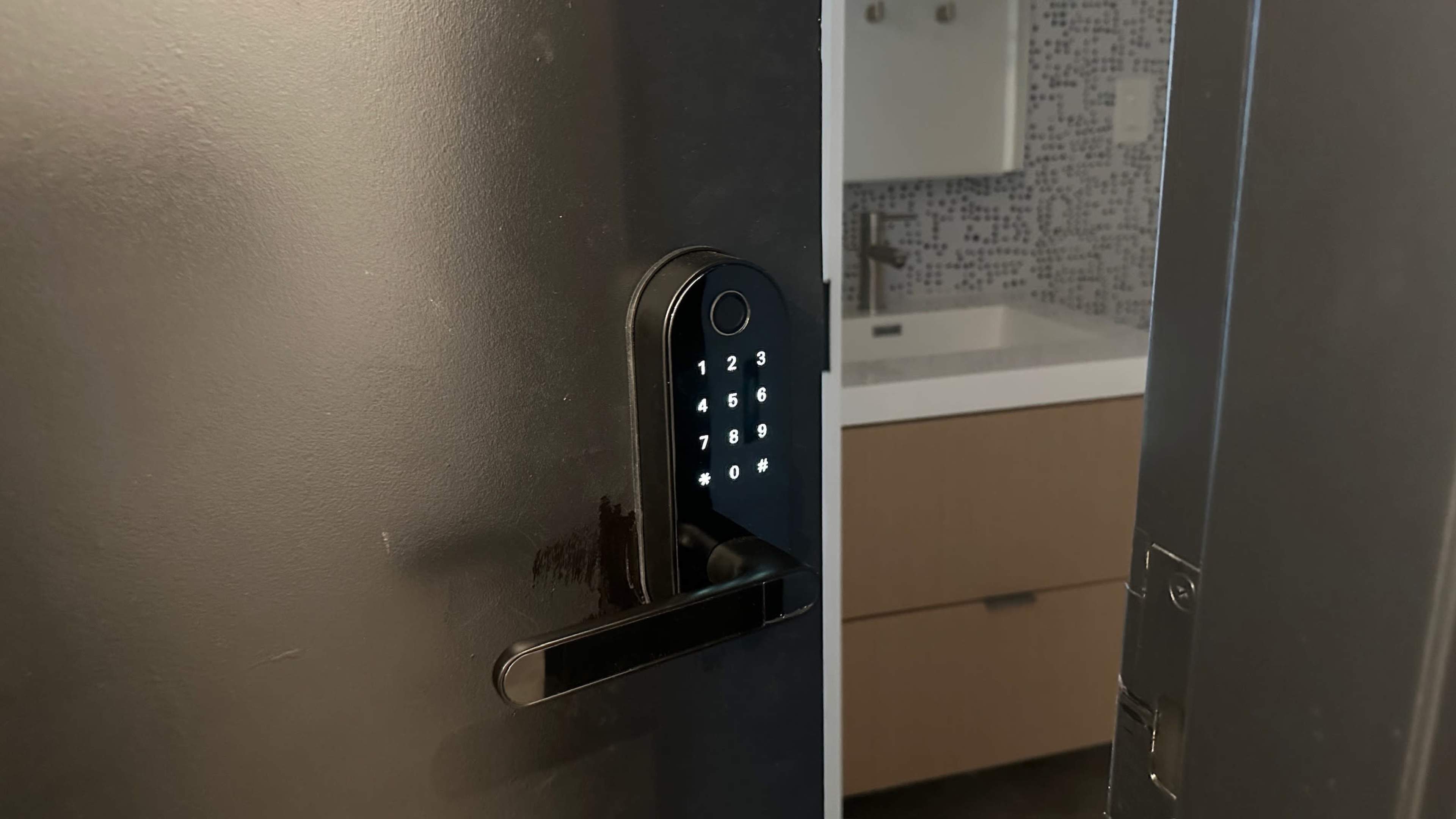 A modern keypad door lock is installed on a sleek black door, with a bathroom visible in the background featuring a countertop and patterned tiles.