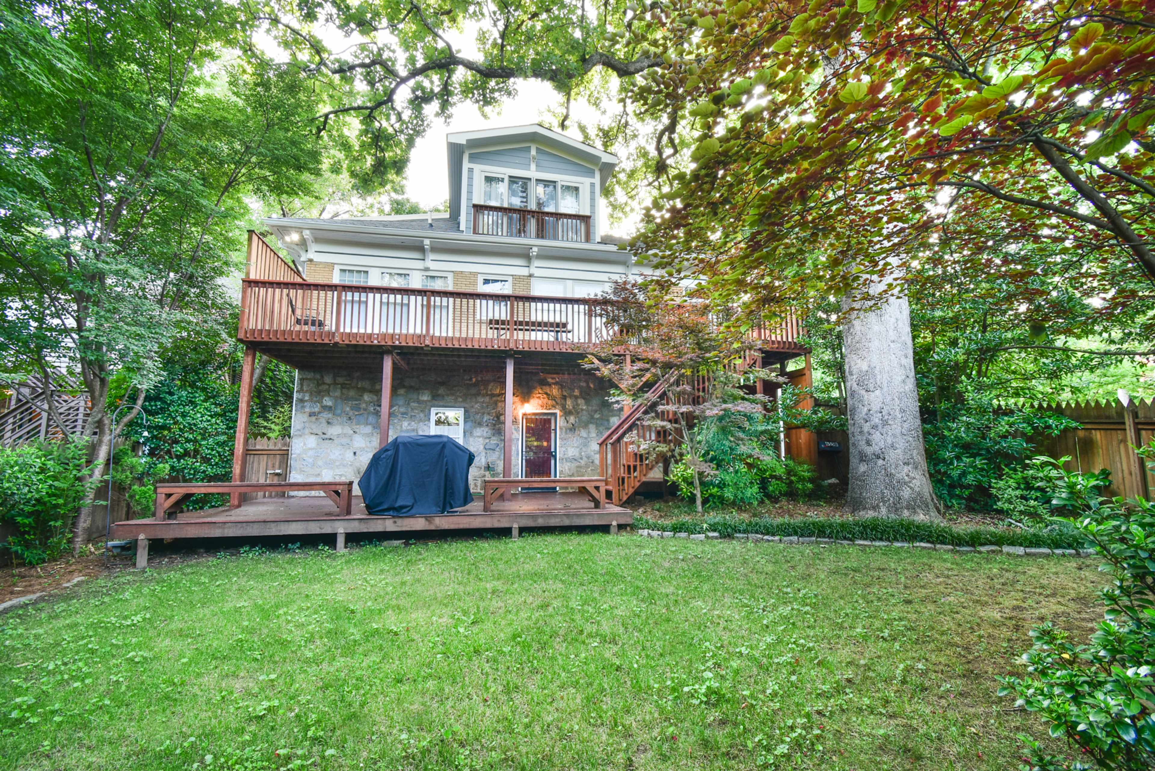 A multi-level house with a large wooden deck in the backyard, surrounded by trees and greenery.