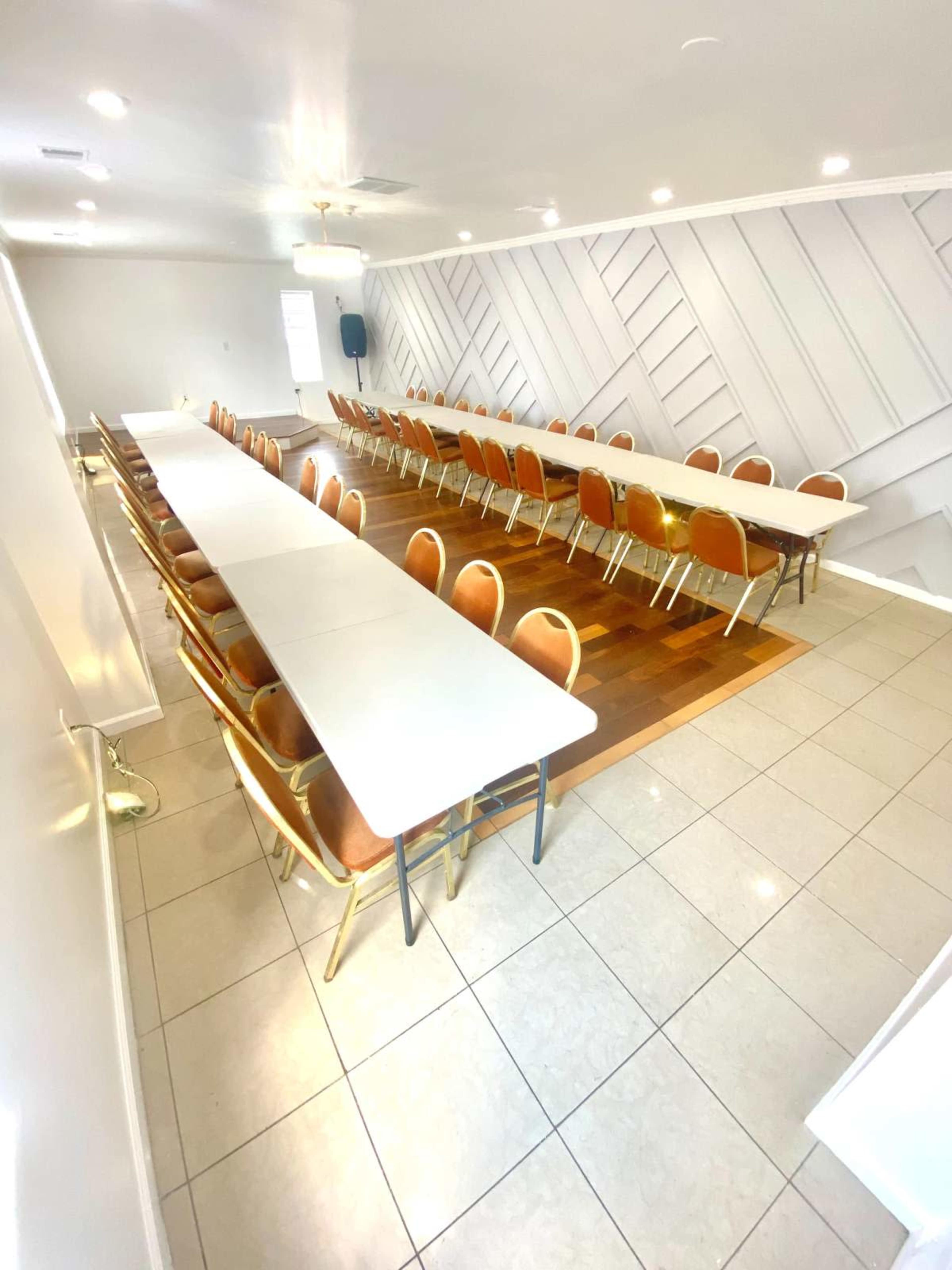 A spacious meeting room features multiple long tables arranged in a U-shape with brown chairs and a tiled floor.