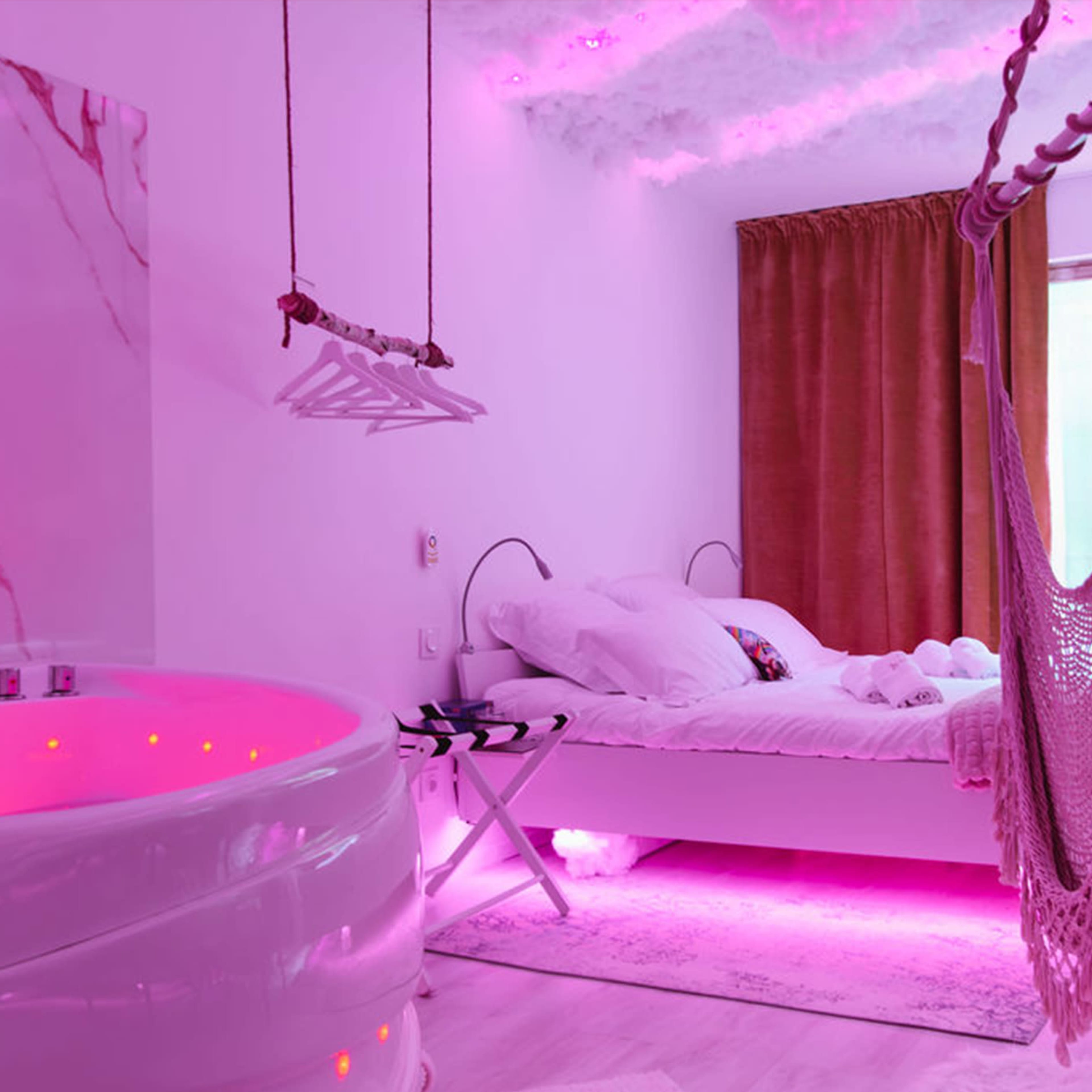 The image shows a modern bedroom featuring a round bathtub with pink lighting, a bed with white linens, and a hammock hanging in the corner.