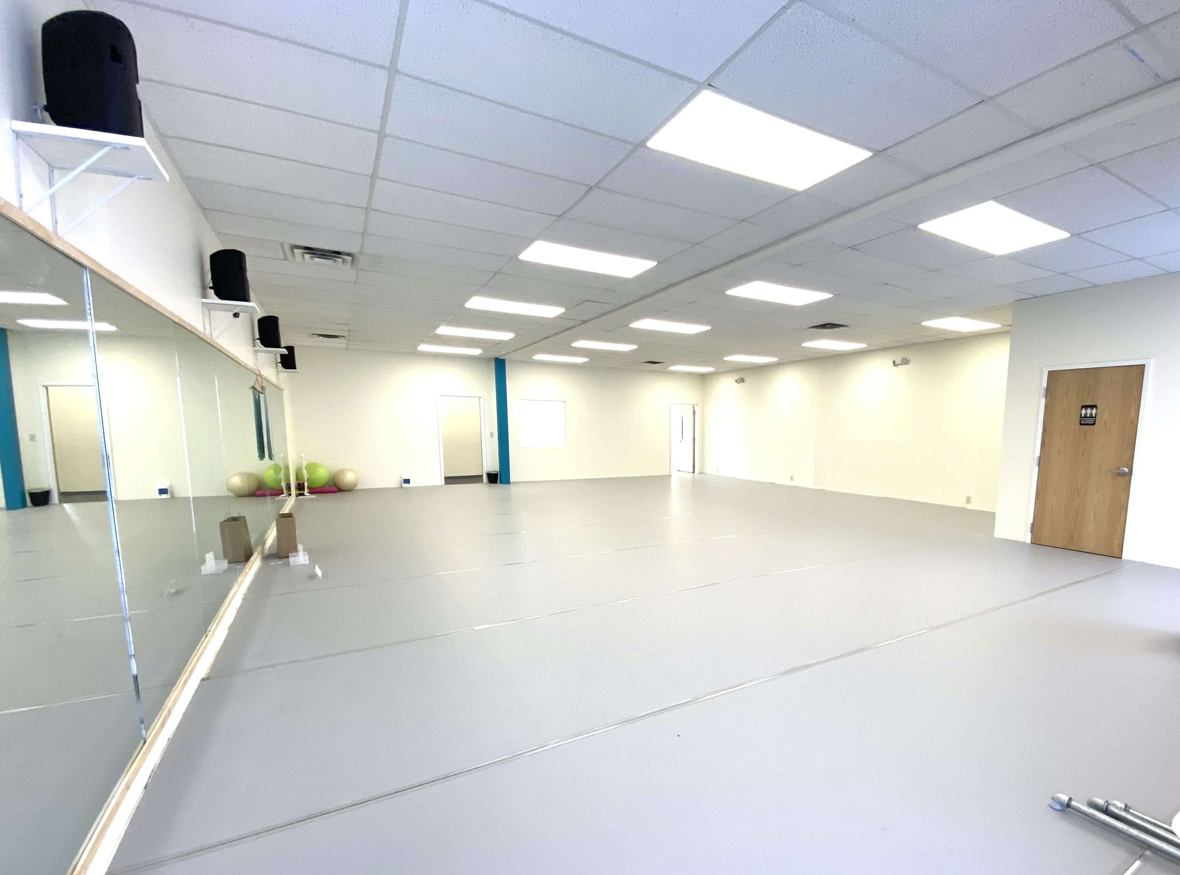 A spacious, empty room with a gray floor, illuminated by overhead lights, featuring a large mirror on one wall and a door on the opposite side.