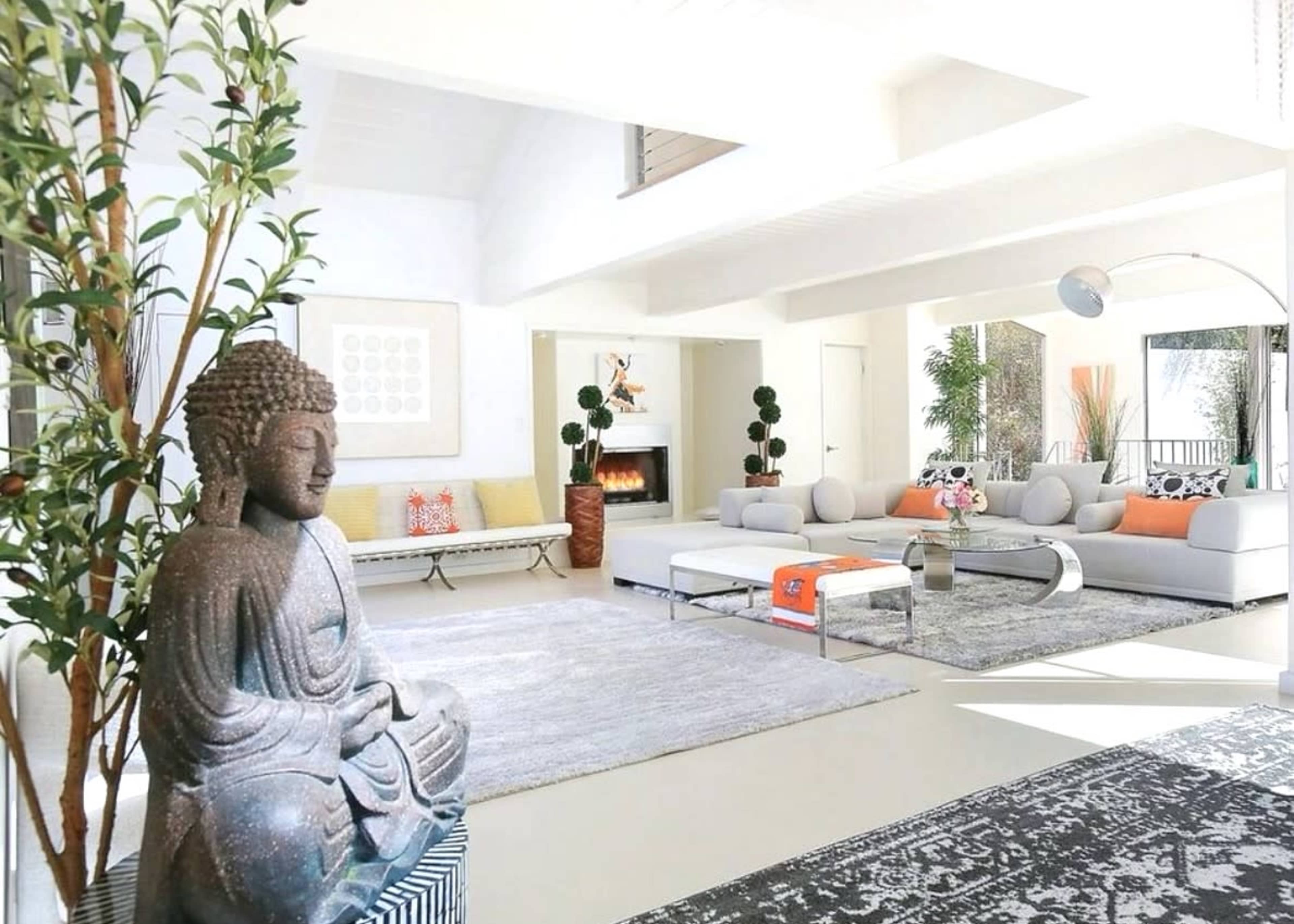 The scene features a bright and airy living room with a large Buddha statue, modern furniture, potted plants, and a fireplace.