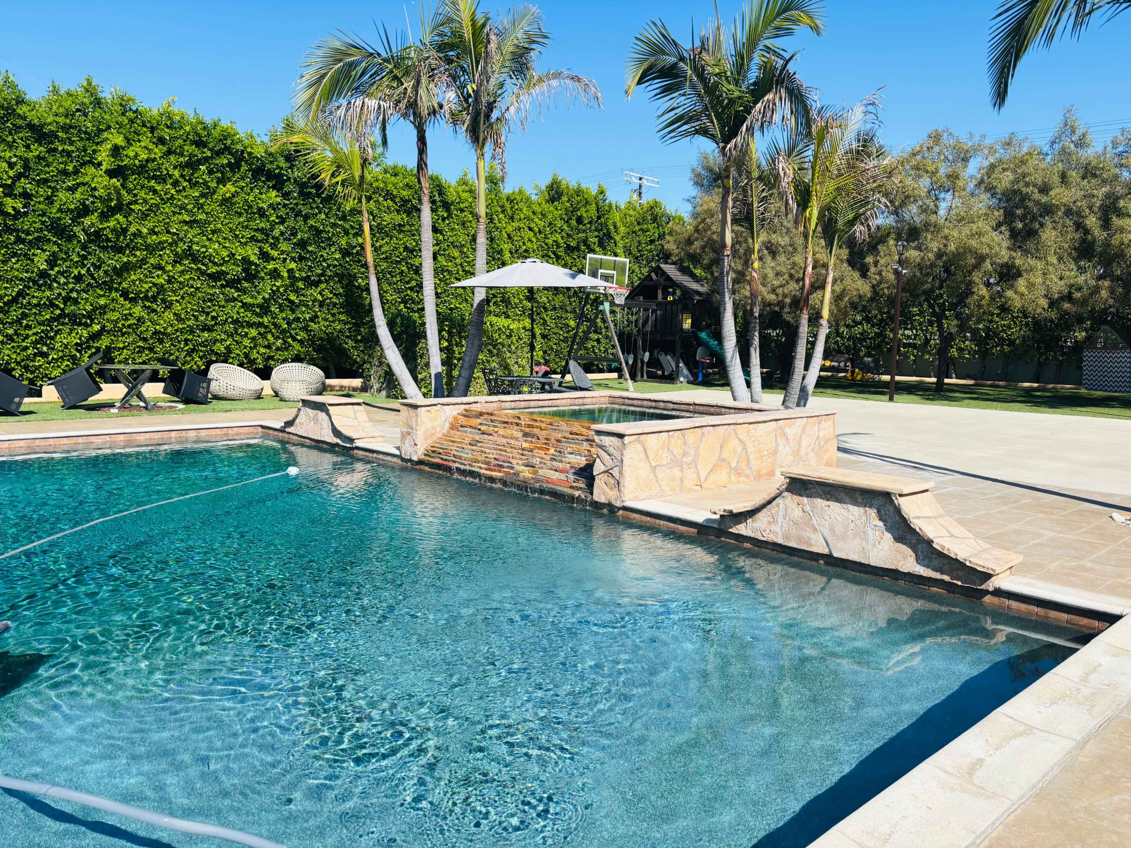 The image shows a backyard pool surrounded by lush greenery, with lounge chairs, palm trees, and a shaded seating area nearby.