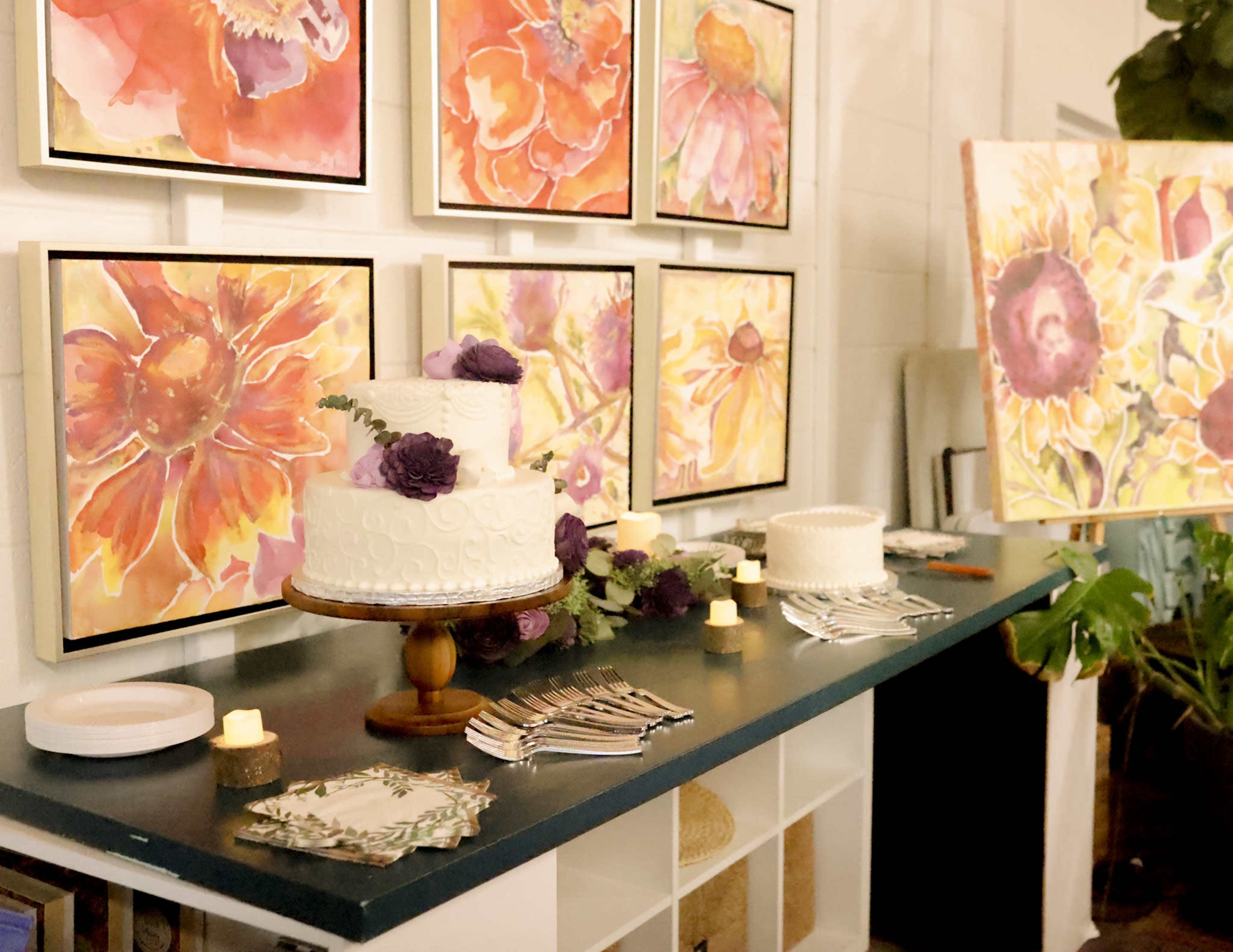 A decorated table displays two wedding cakes, colorful flower paintings on the wall, and other event setup items.