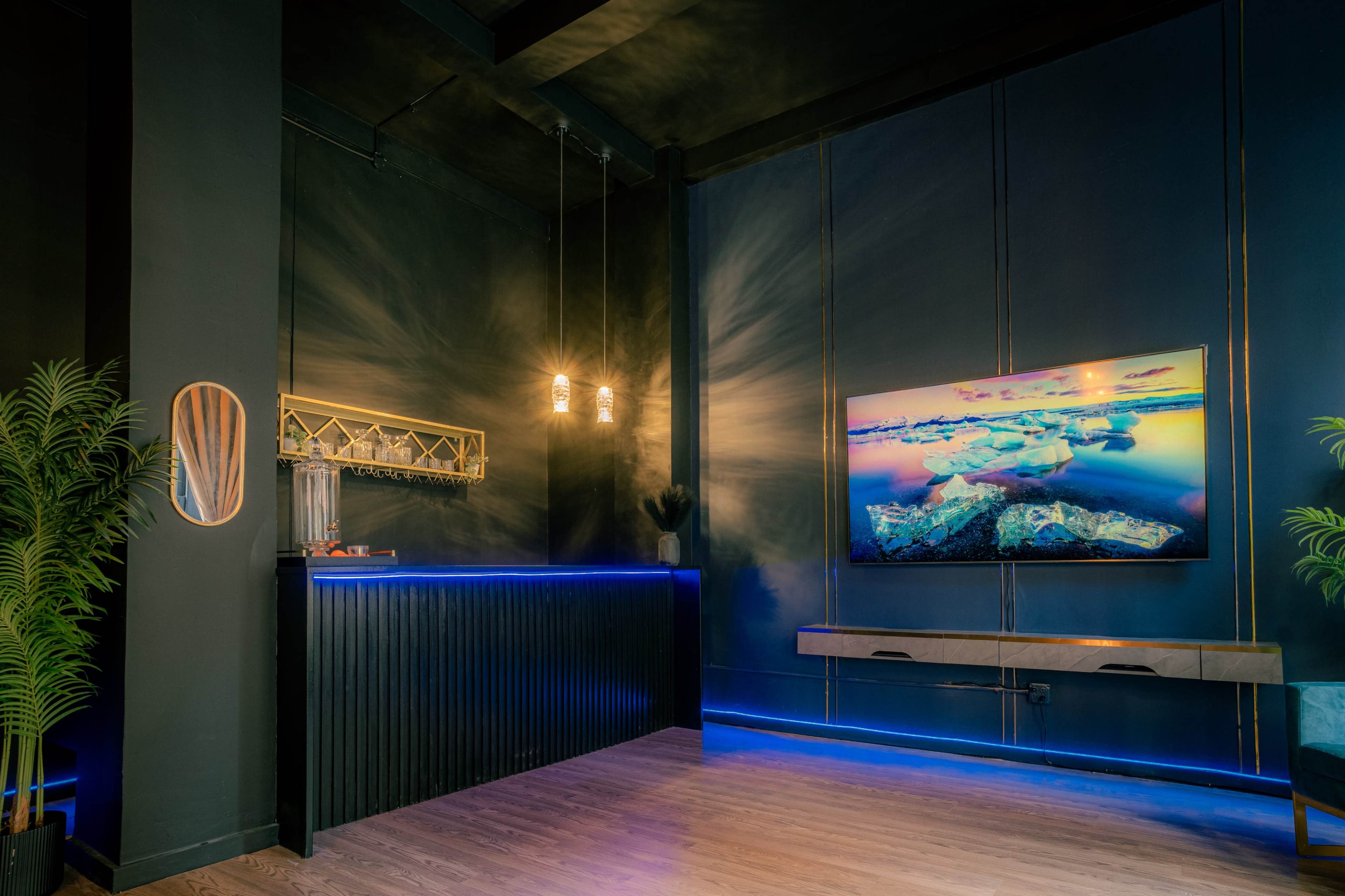 A modern reception area with a dark color scheme, featuring a bar counter, a wall-mounted television displaying landscape imagery, and decorative plants.
