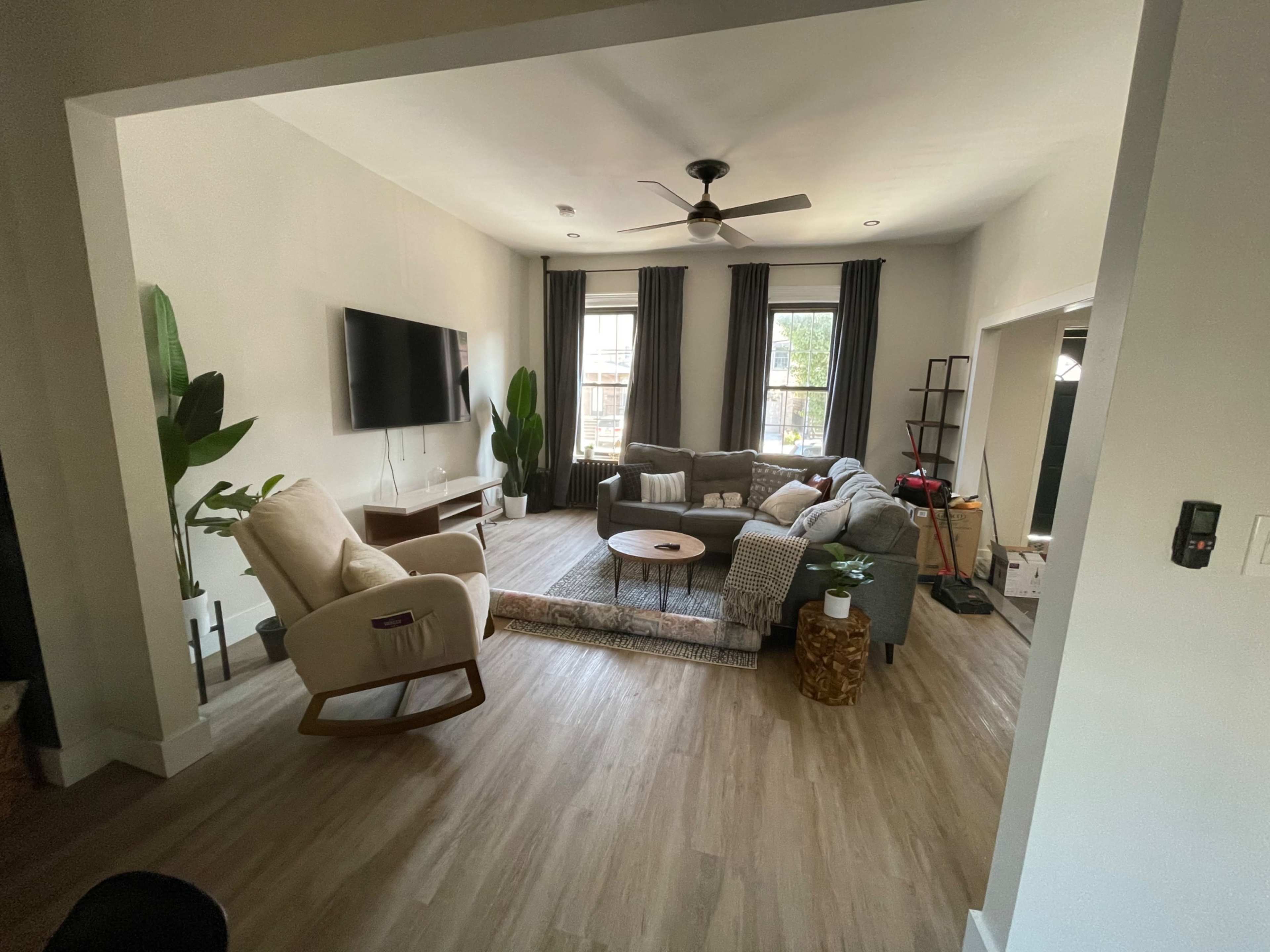 A living room with a gray sectional sofa, a round coffee table, a television mounted on the wall, and large windows with curtains.