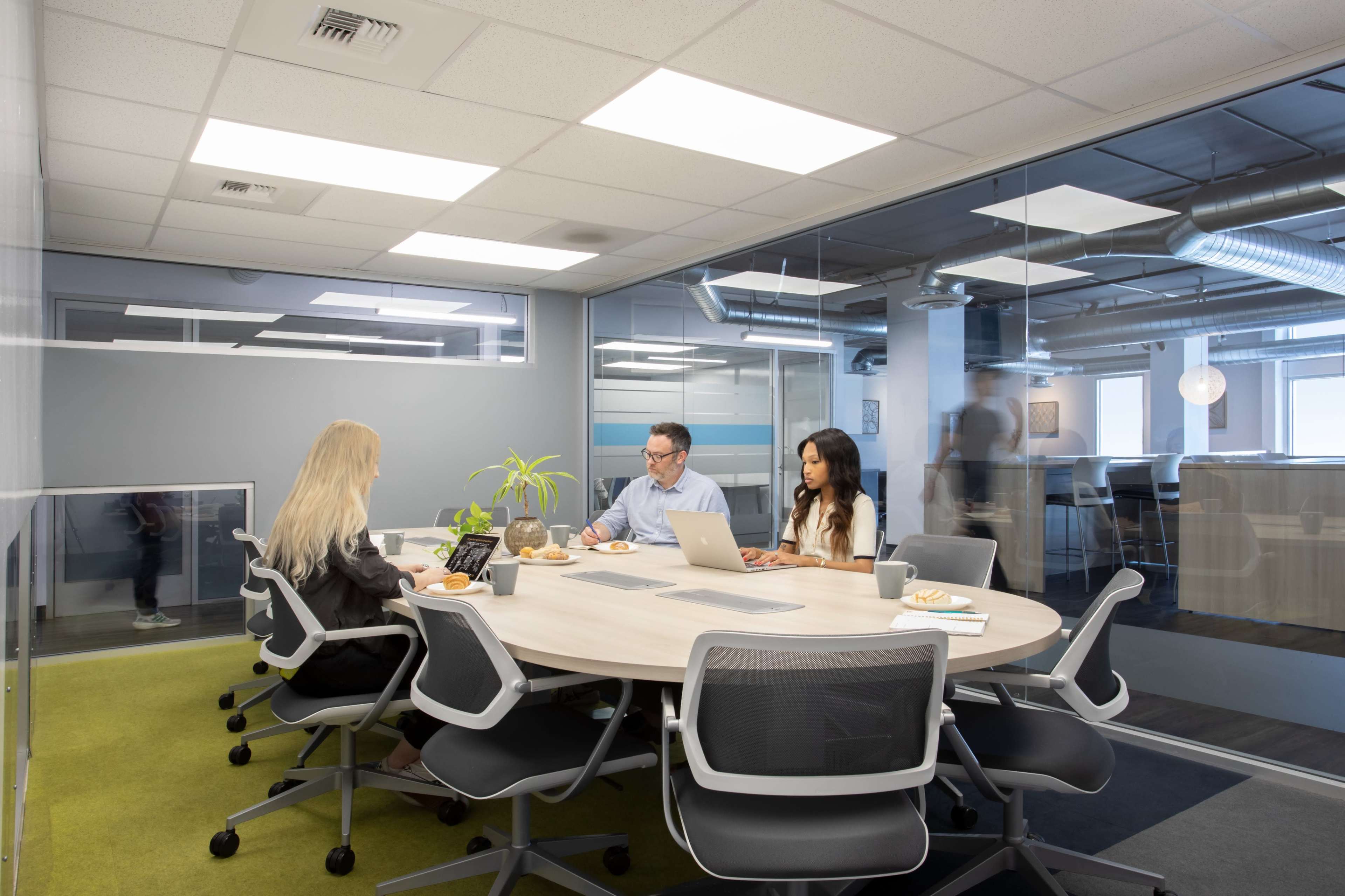 A meeting room features three individuals engaged in discussion around a round table, with a plant and laptops present on the table.