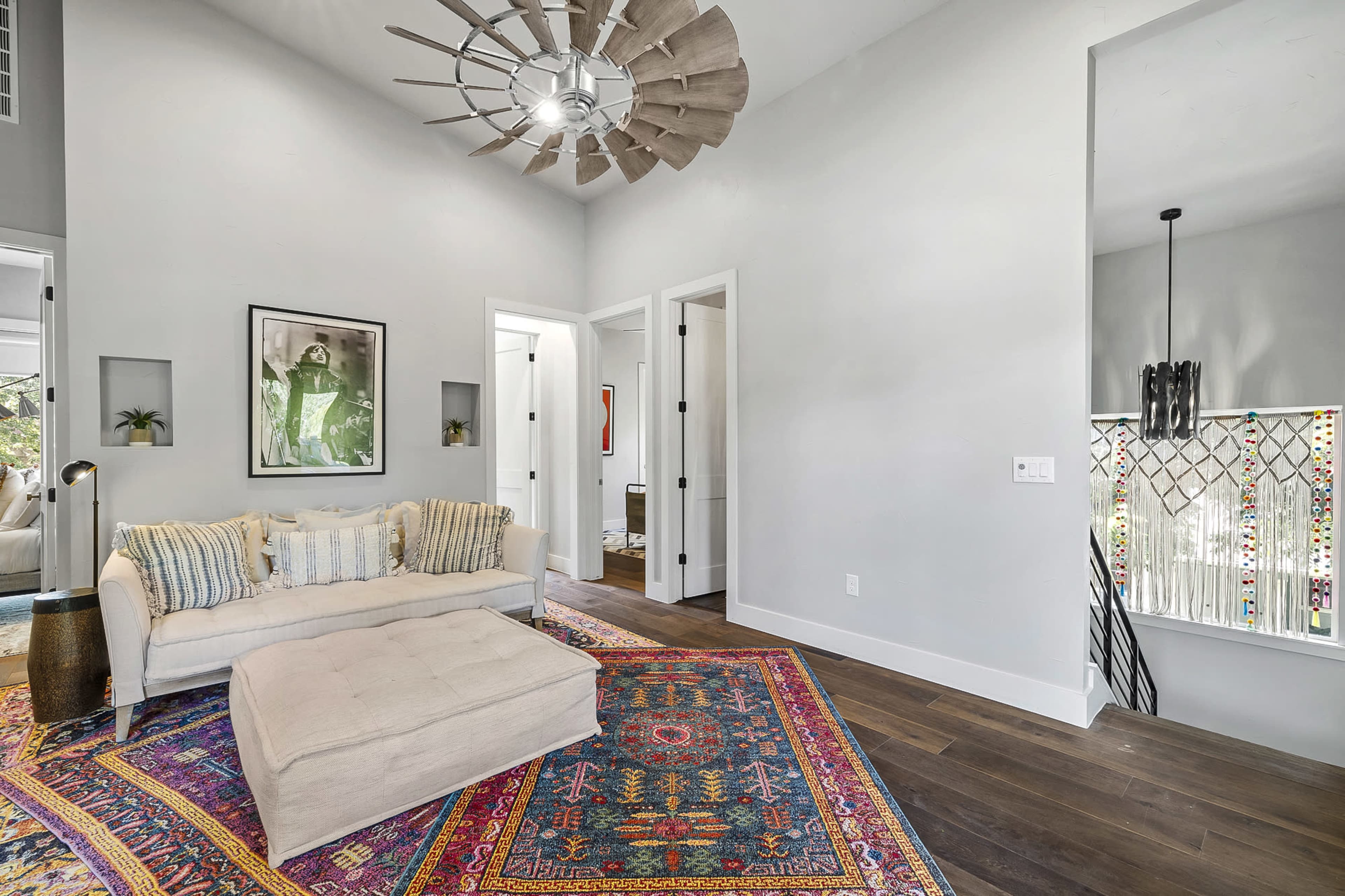 The image displays a modern living room with a neutral-colored couch on a colorful patterned rug, a large ceiling fan, and brightly lit walls.