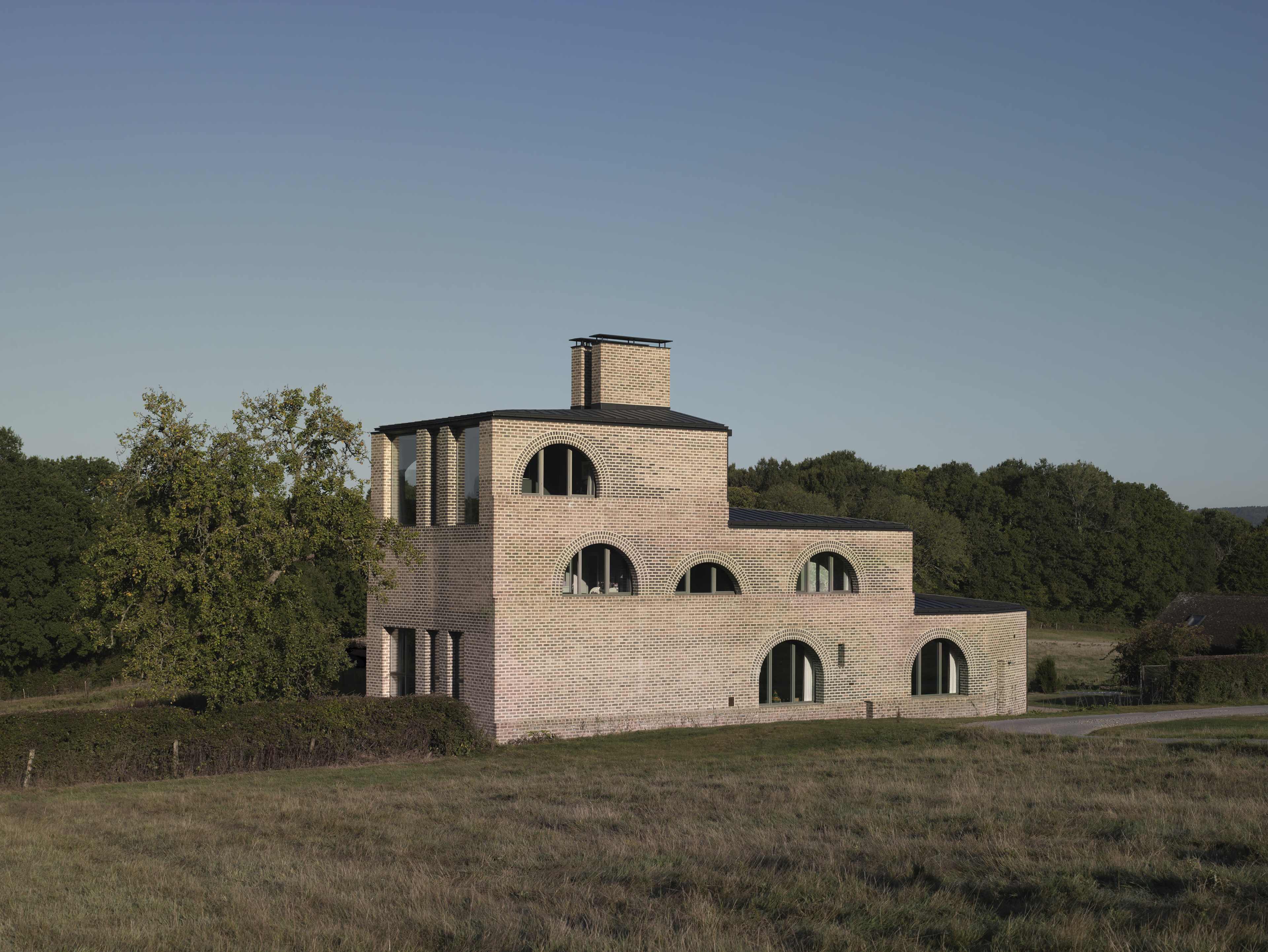 A modern, multi-level brick house with arched windows sits on a grassy landscape surrounded by trees.