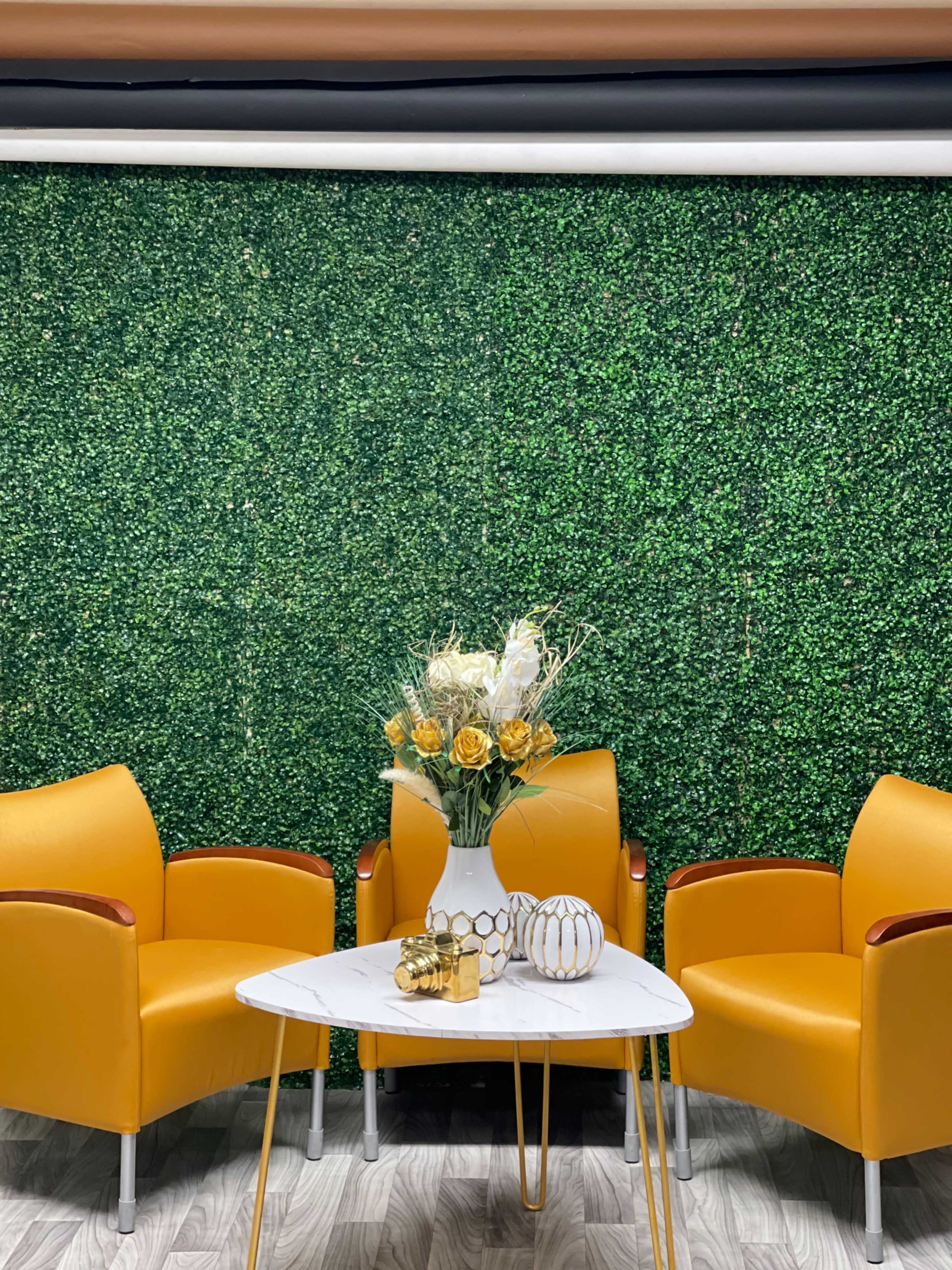 Two yellow armchairs facing a white marble table, with a vase of flowers on the table, set against a green artificial hedge background.