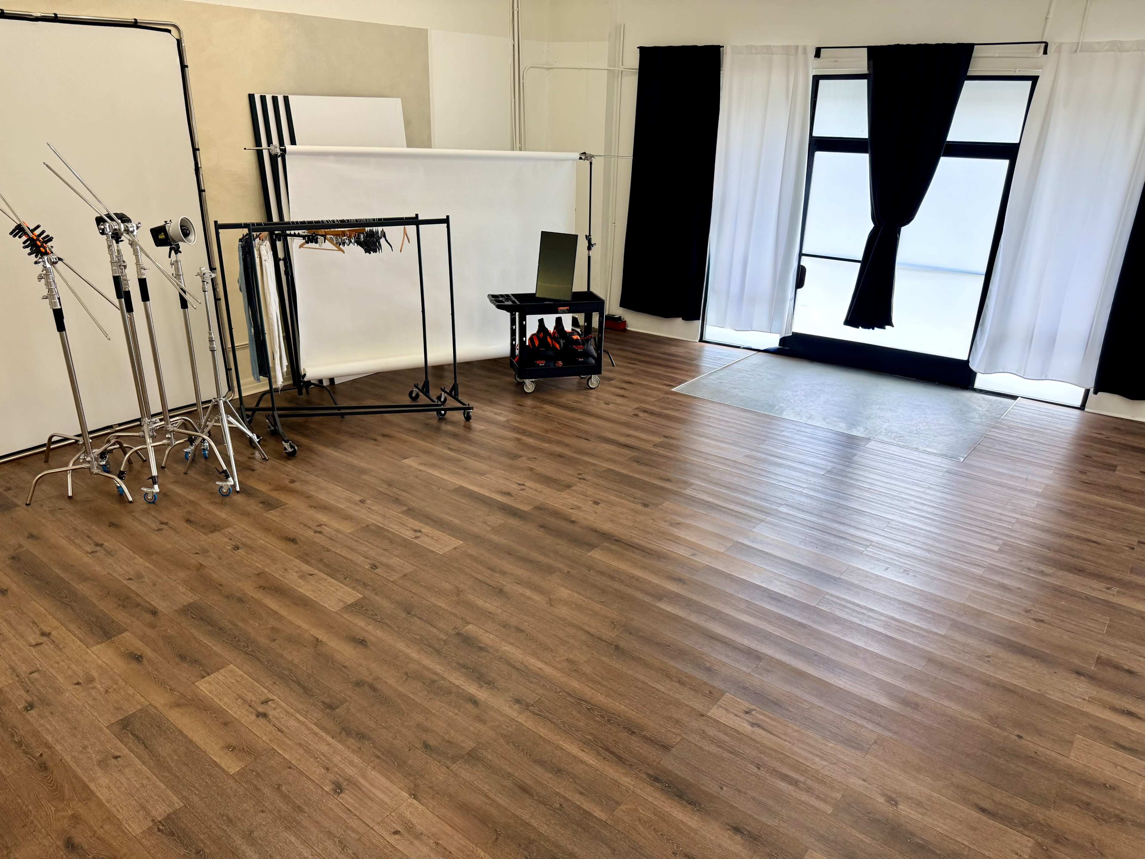 The image shows a spacious, empty studio room with wooden flooring, featuring a backdrop stand, a cart with equipment, and large windows covered by black curtains.