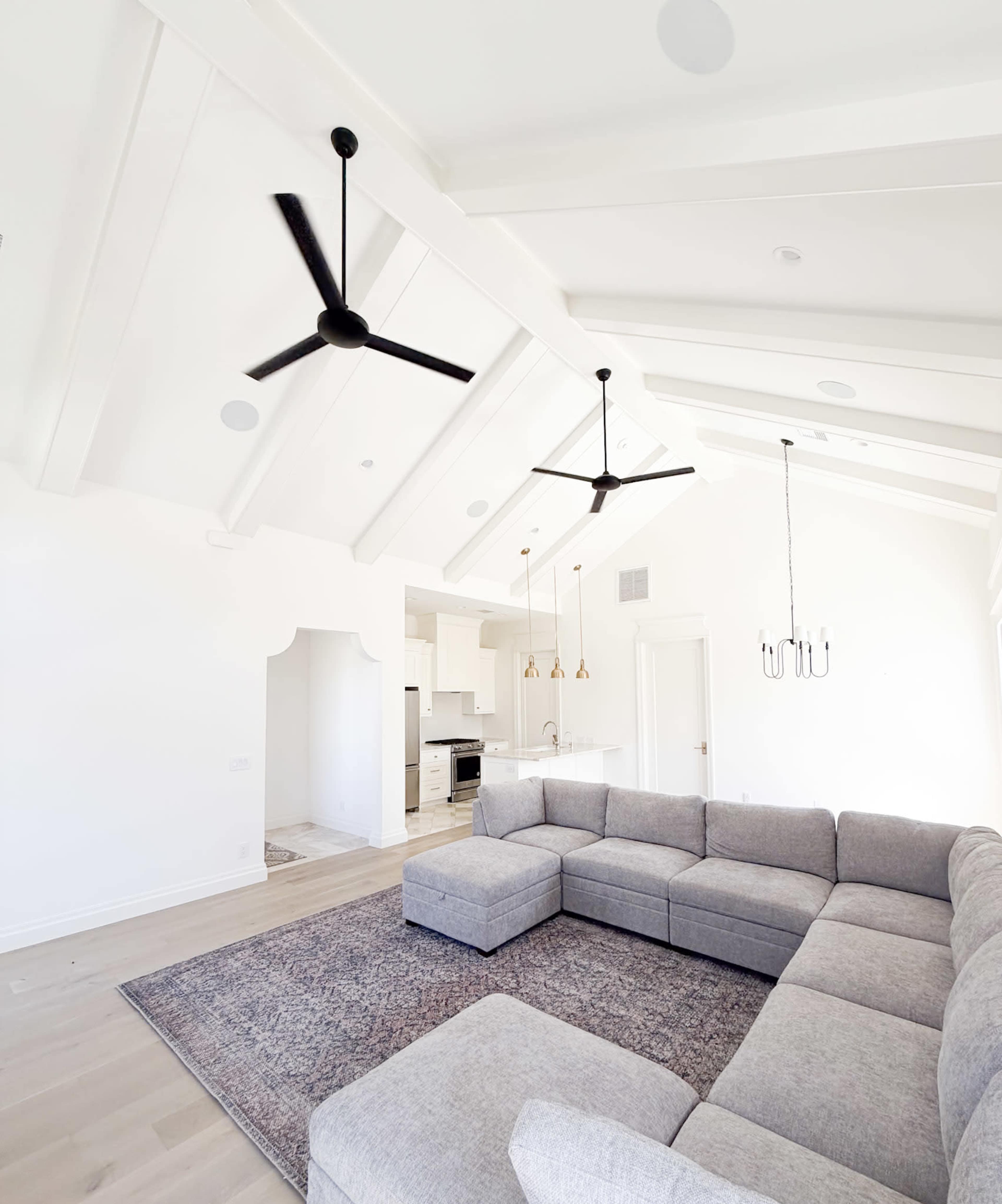 A modern living room features a large gray sectional sofa, two ceiling fans, and a kitchen area visible in the background.