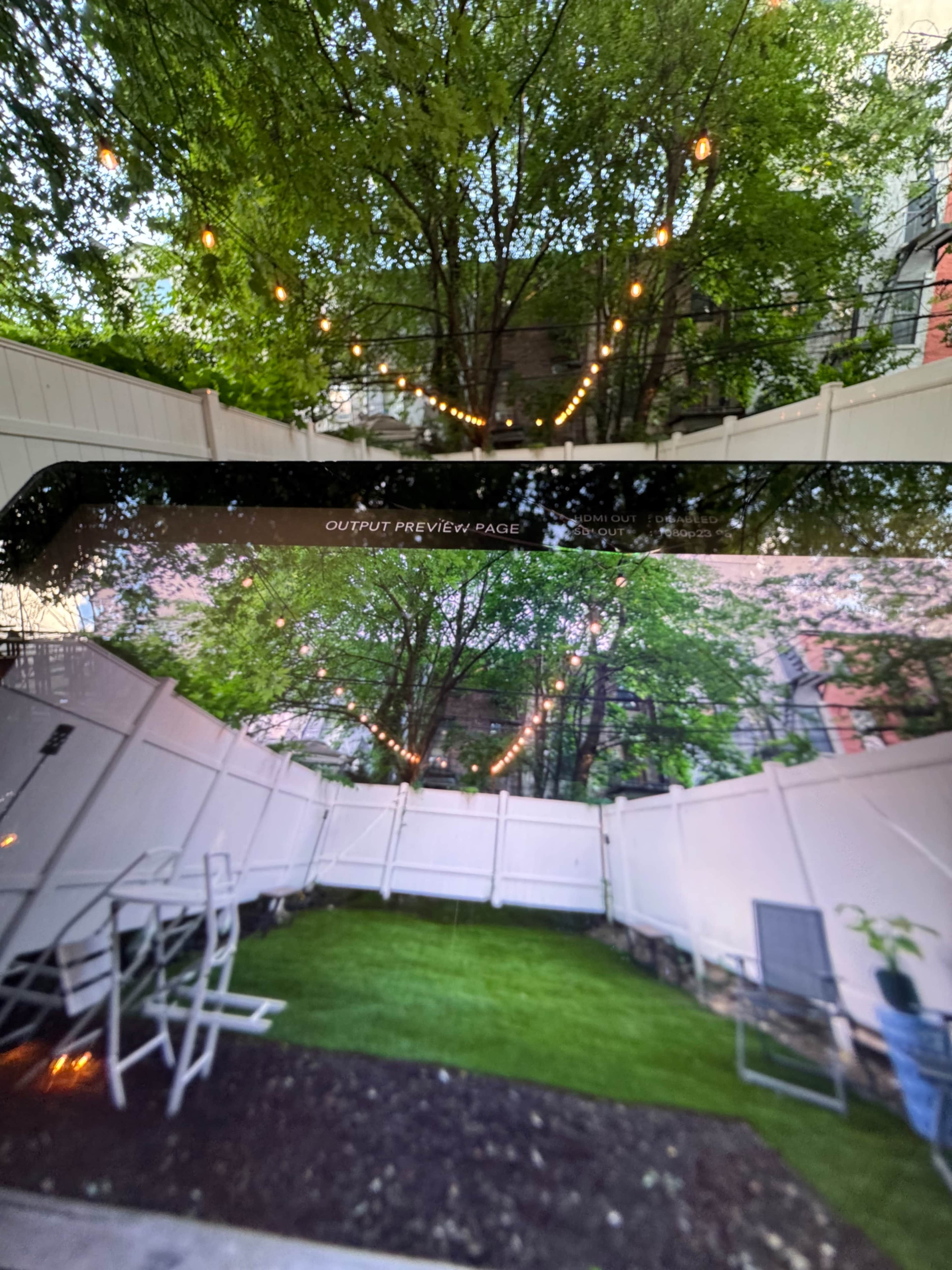 A backyard scene with a grassy area surrounded by a white fence, adorned with string lights hanging from the trees above.