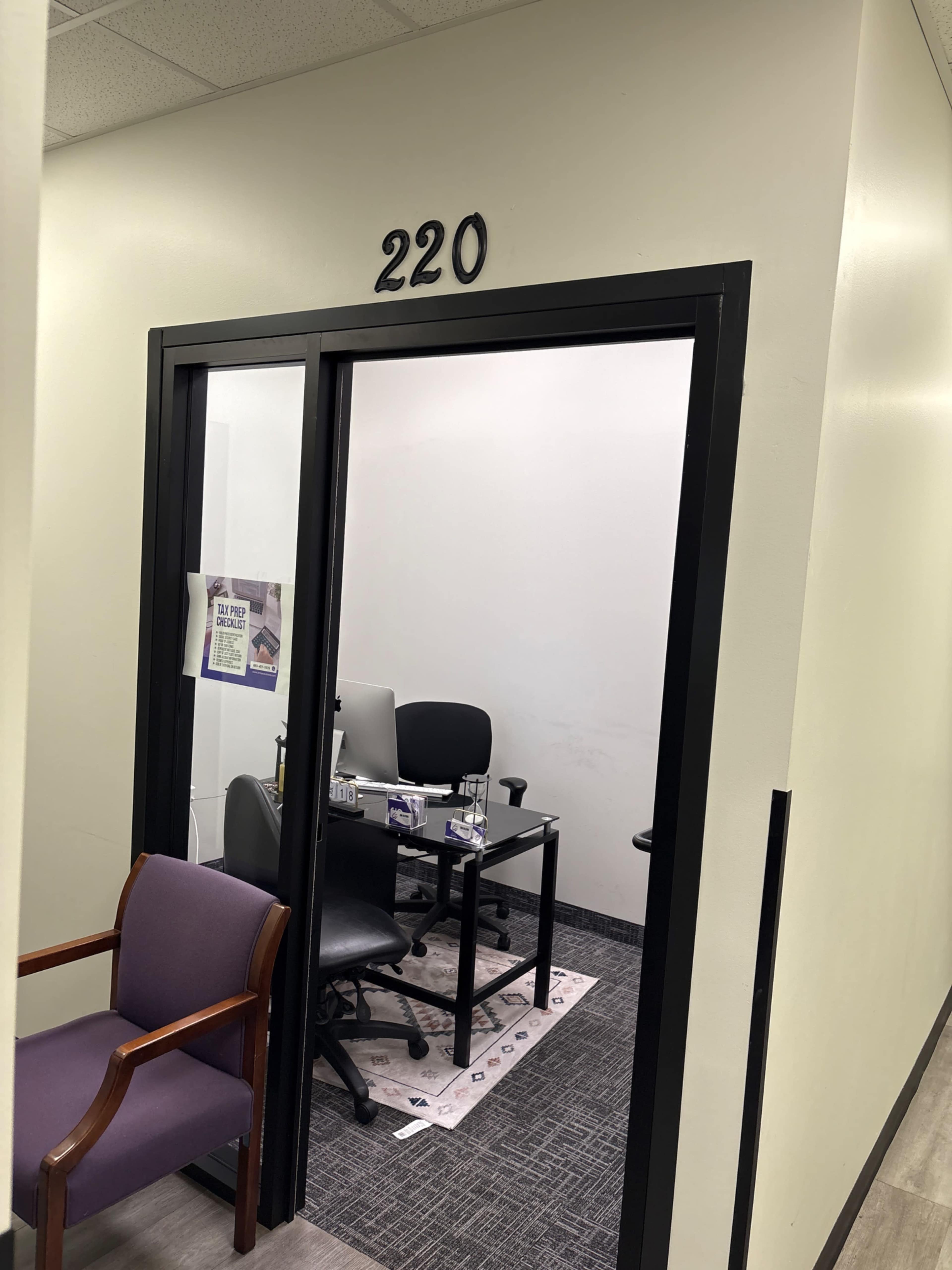 The image shows the entrance to office number 220, featuring a glass door that opens into a small meeting room with two chairs and a table.