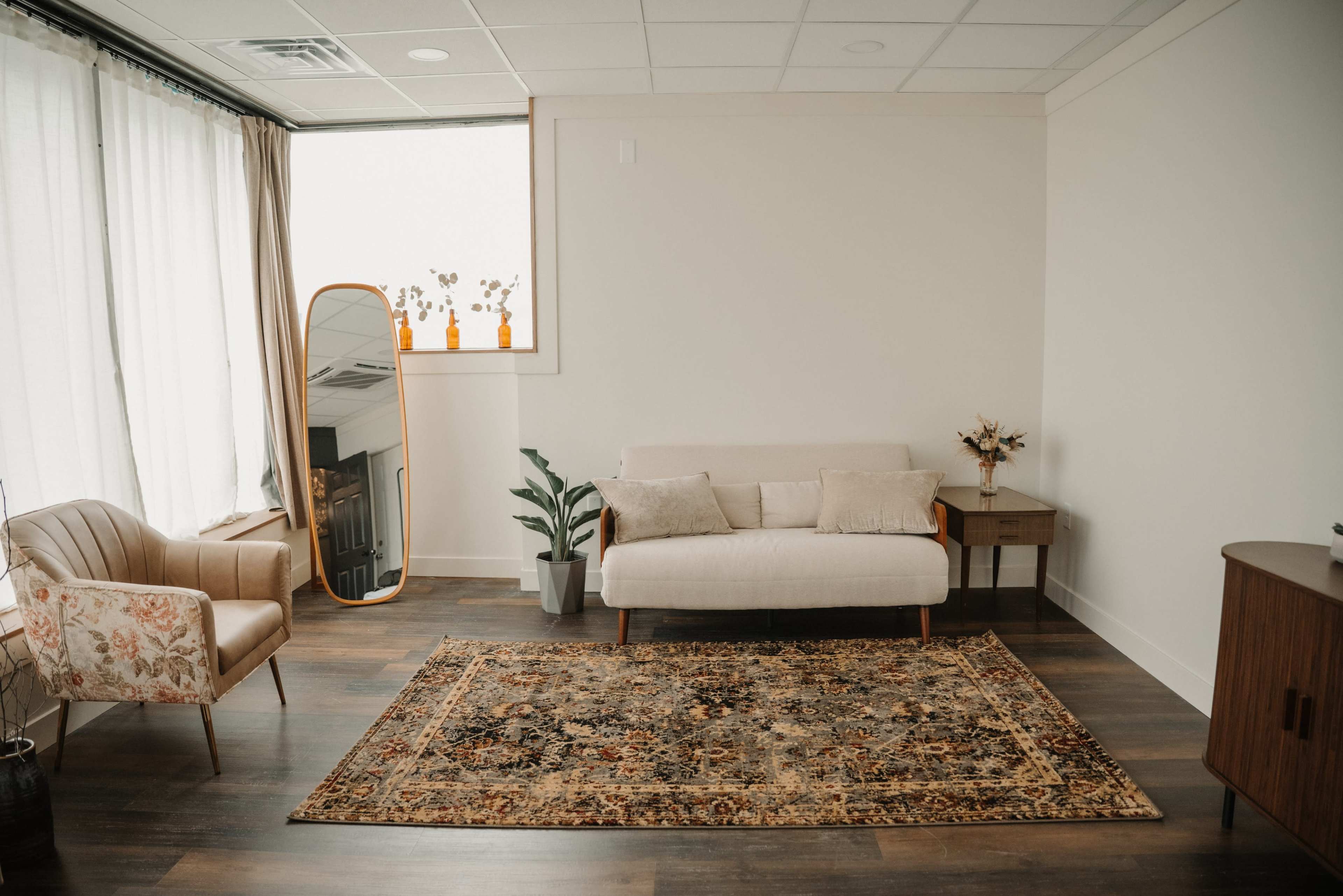 The image features a bright, modern room with a sofa, a floral armchair, a decorative rug, a plant, a side table, and a mirror.