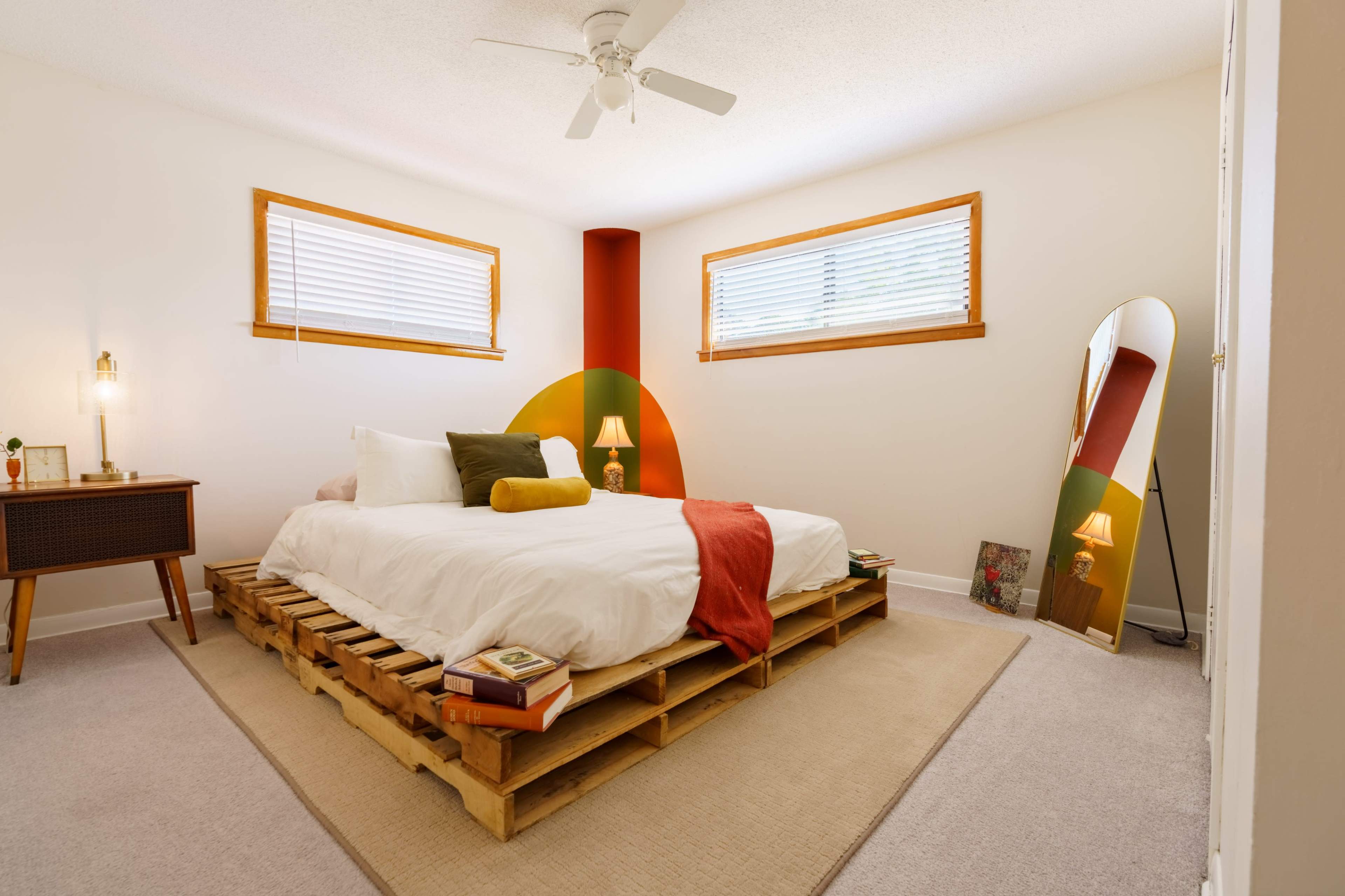 The image shows a cozy bedroom with a bed on a wooden pallet, light-colored walls, and two windows letting in natural light.