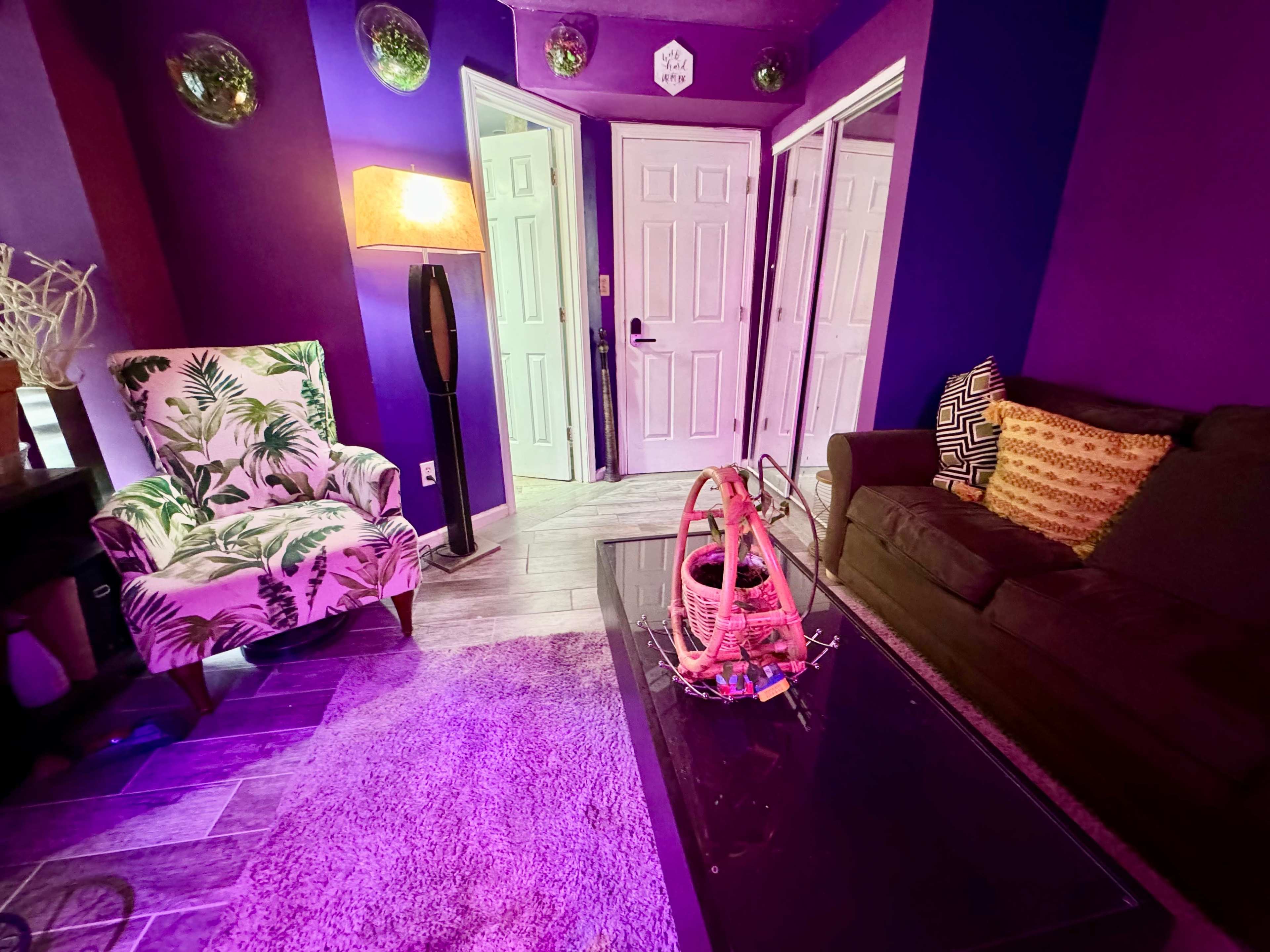 A living room with purple walls, a patterned armchair, a dark couch, and a coffee table in the center, illuminated by a lamp.